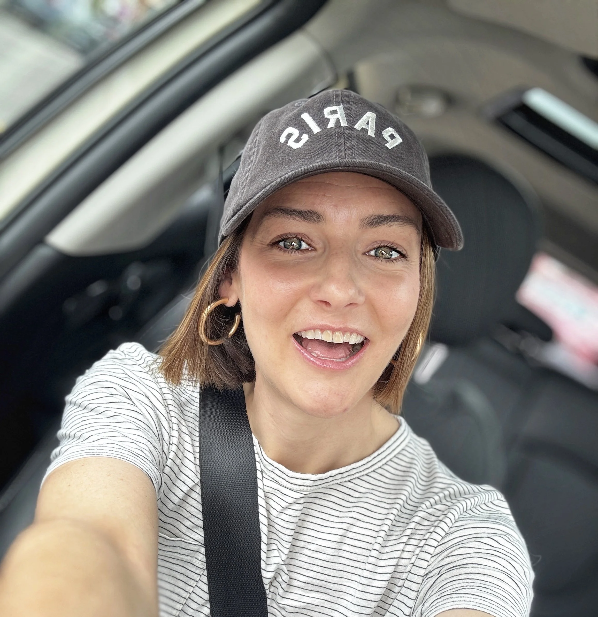 A woman taking a selfie inside a car, wearing a gray baseball cap with the word 'PARIS' embroidered on it, smiling with her mouth open and showing her teeth.