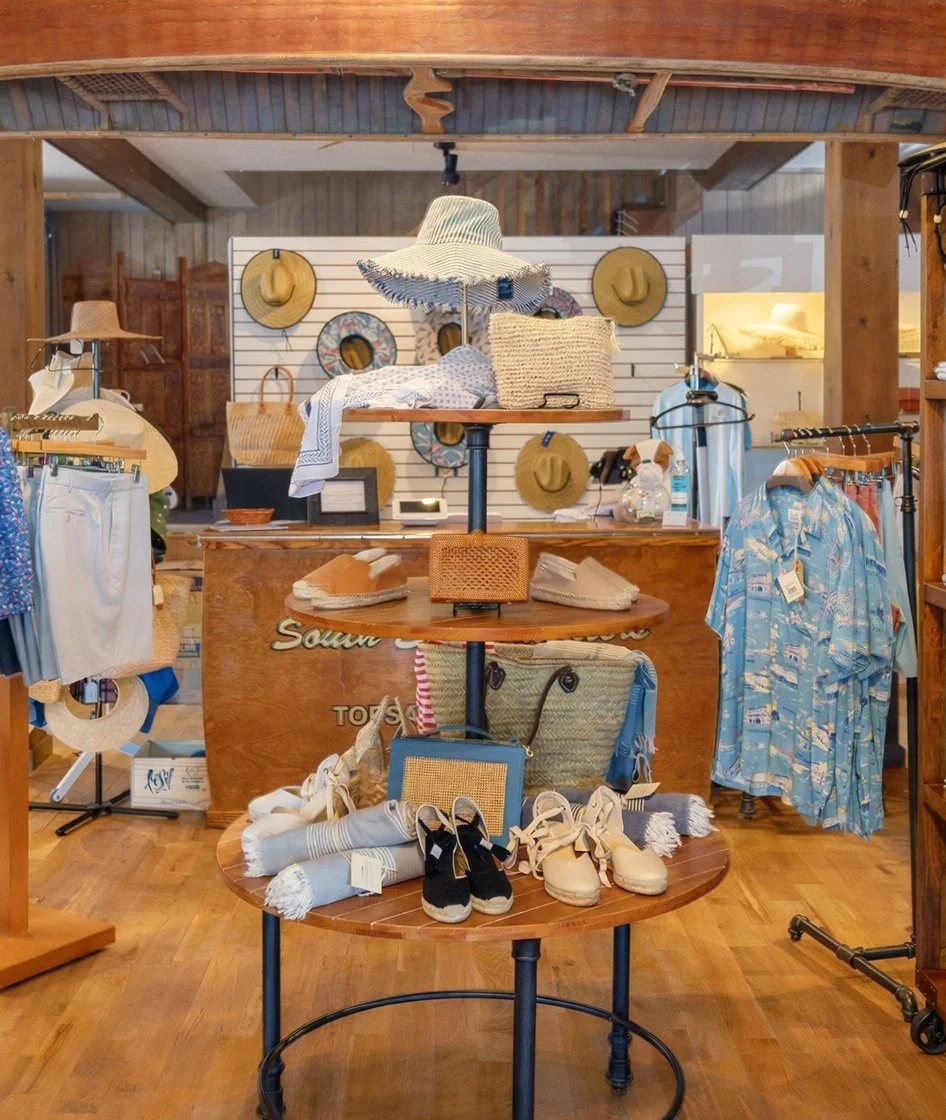 Display of summer accessories and clothing in a retail store, including hats, straw bags, towels, shoes, and a blue Hawaiian shirt.