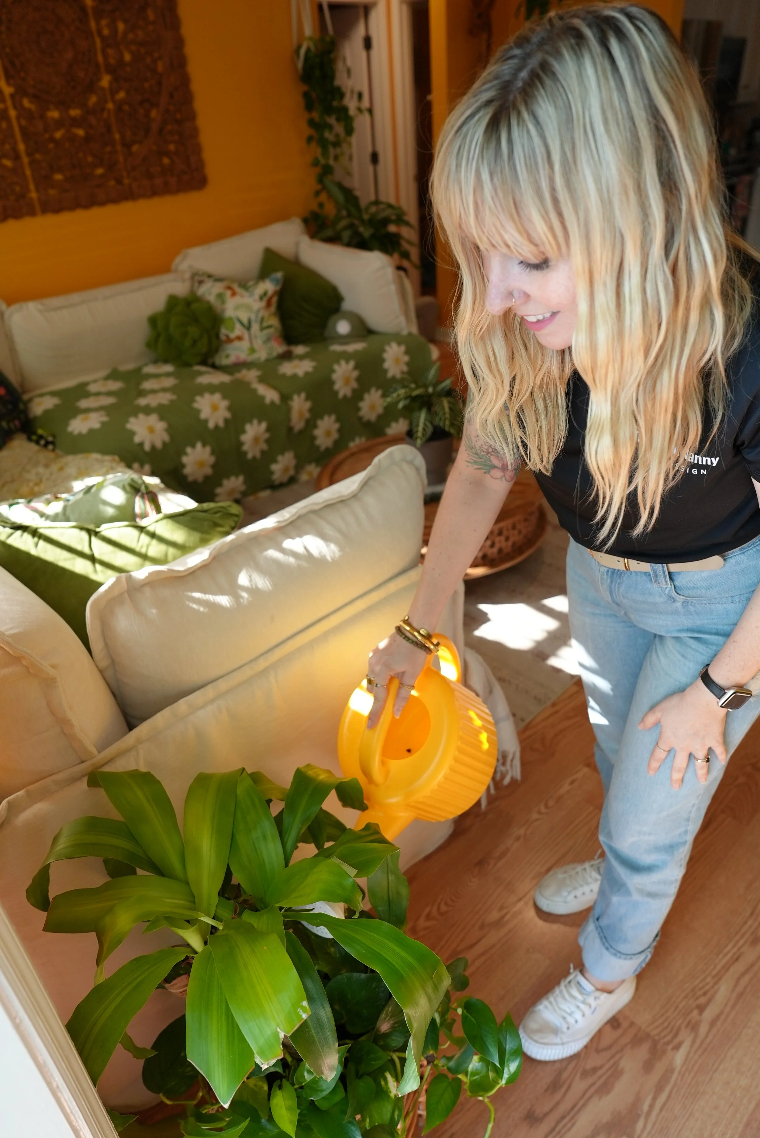 A woman watering houseplants in a cozy living room with colorful cushions and a yellow wall. She is plant sitting for someone who needs their plants watered while they're out of town on vacation. They call her The Plant Nanny!