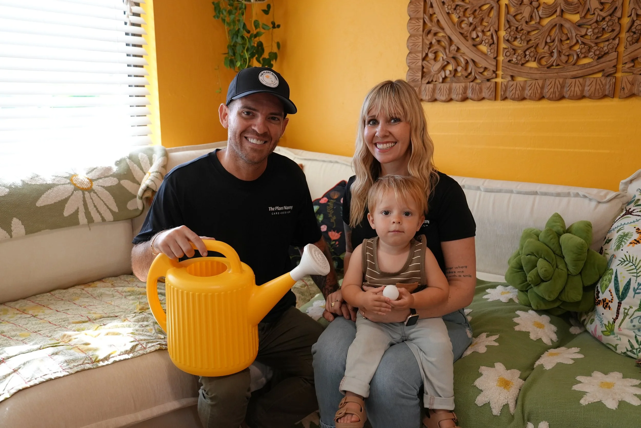 A family of three sitting on a couch in a colorful living room. The man on the left is holding a yellow watering can, the woman on the right has her arm around a young child sitting on her lap. This is the family that owns, The Plant Nanny home watch