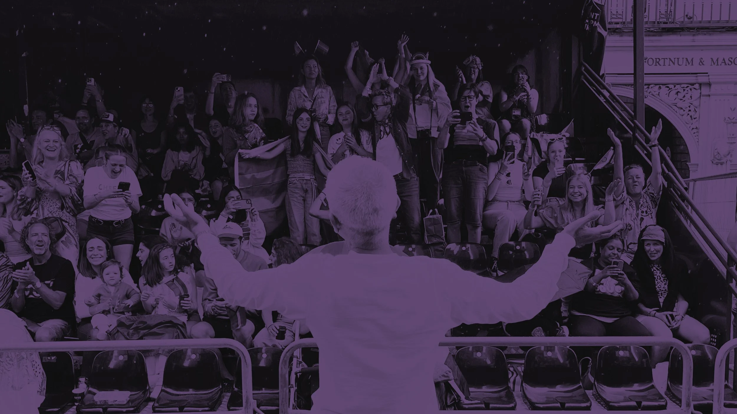 A view from behind Mayor Sadiq Khan as he stands with open arms before a cheering crowd in grandstands. Spectators wave rainbow flags and hold up smartphones at the Pride event.