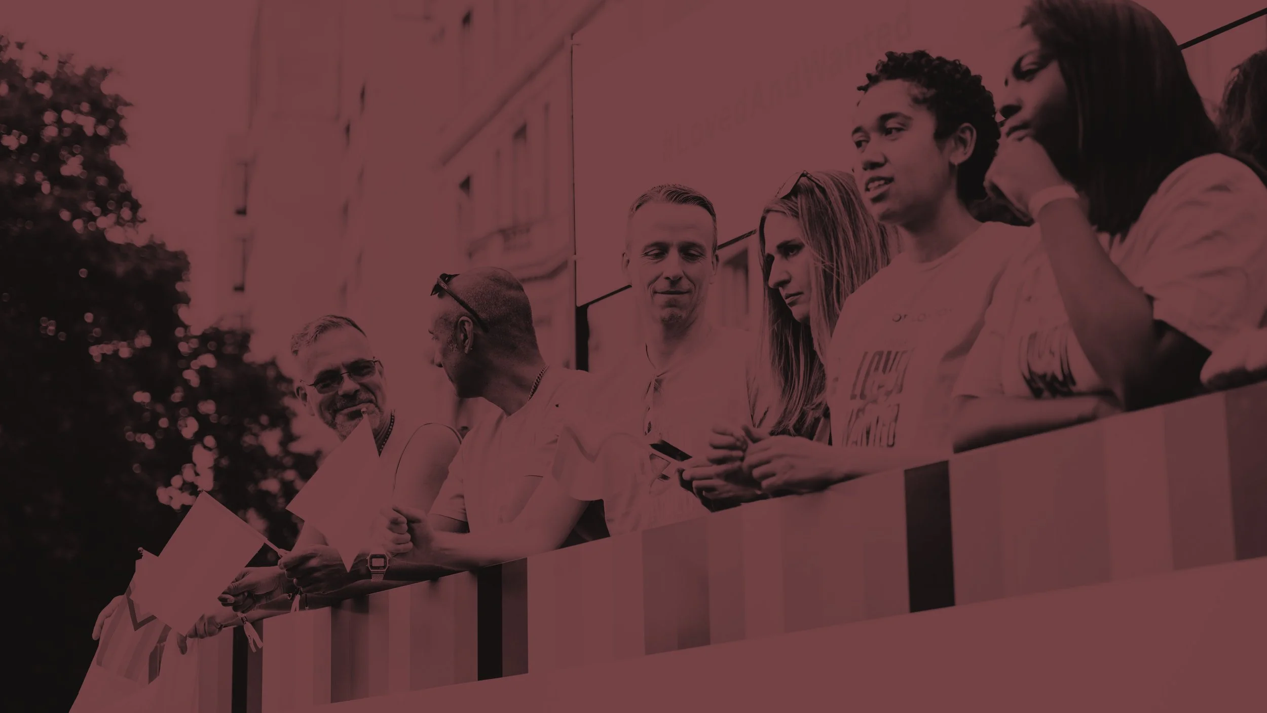 A diverse group of five people, wearing branded "PRIDE OF LONDON" t-shirts and holding flags, stands together on a parade float, looking outwards. A banner above them features text and logo elements.
