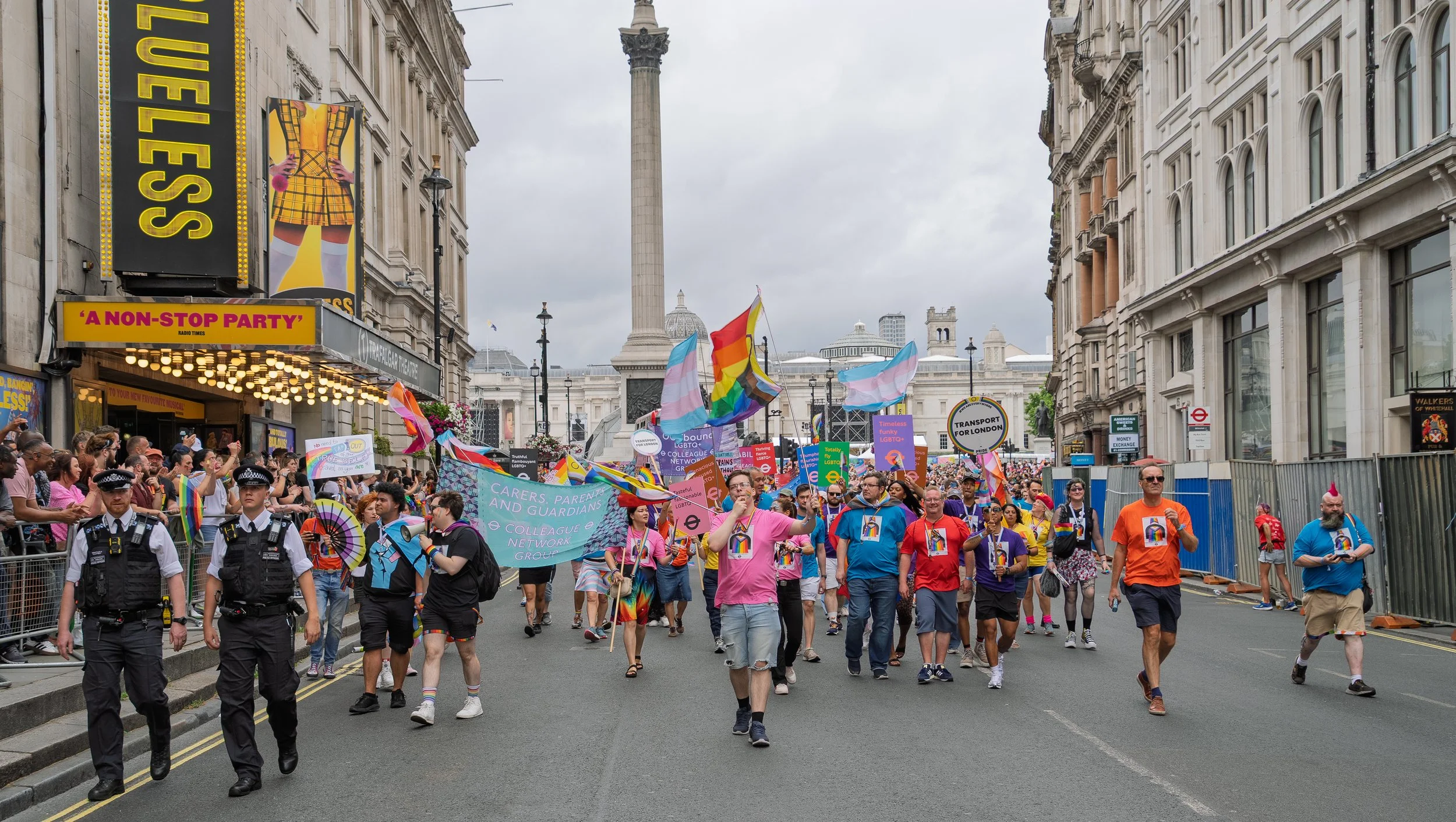 A large, colorful Pride parade marches down a London street, away from Trafalgar Square, with Nelson's Column in the background. Participants are carrying Progress Pride and Transgender flags. Spectators line the pavements under cloudy skies.