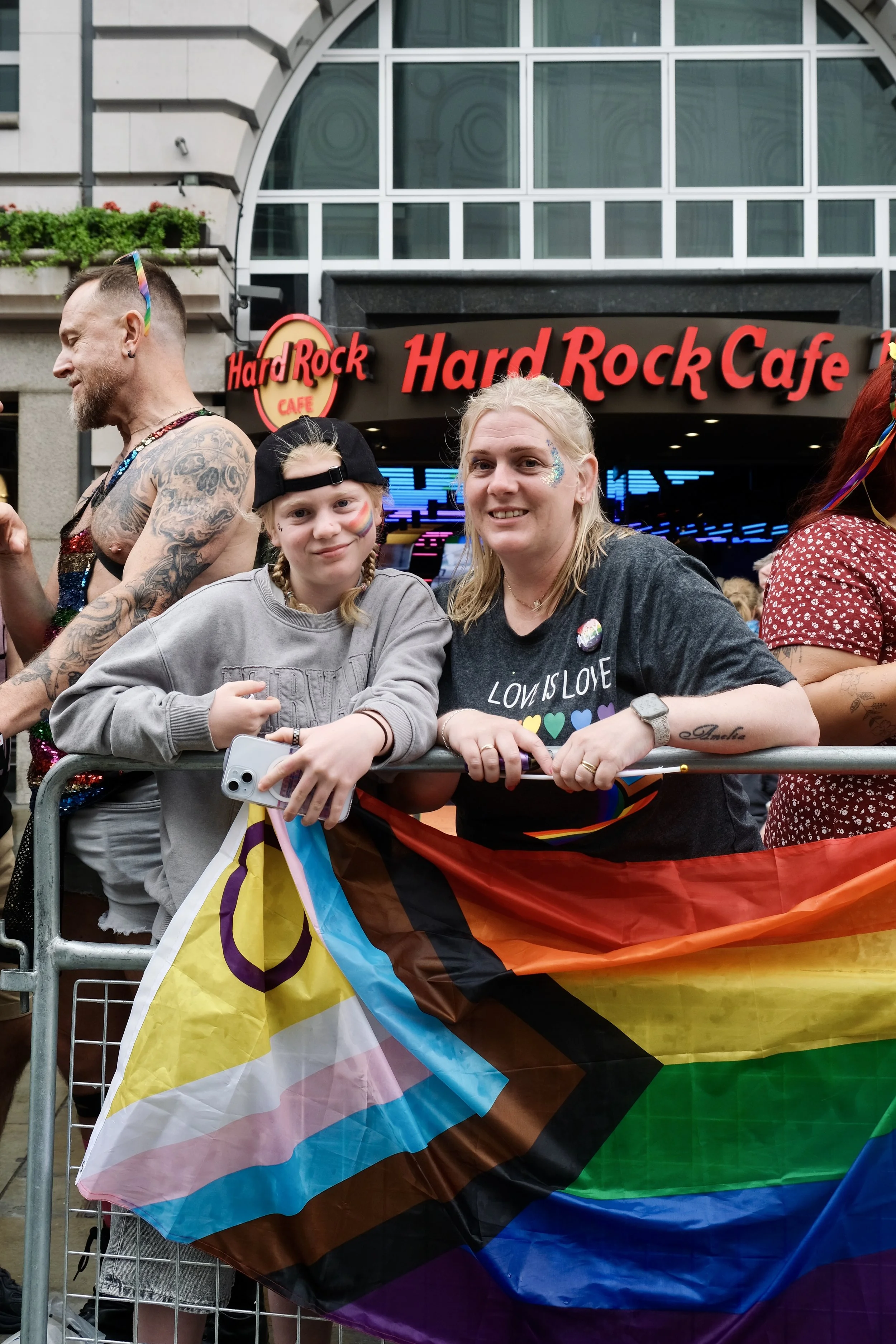 wo attendees, an adult and a young person, lean against a metal crowd barrier during the Pride in London parade. A large Intersex-Inclusive Progress Pride flag is draped over the barrier in front of them.