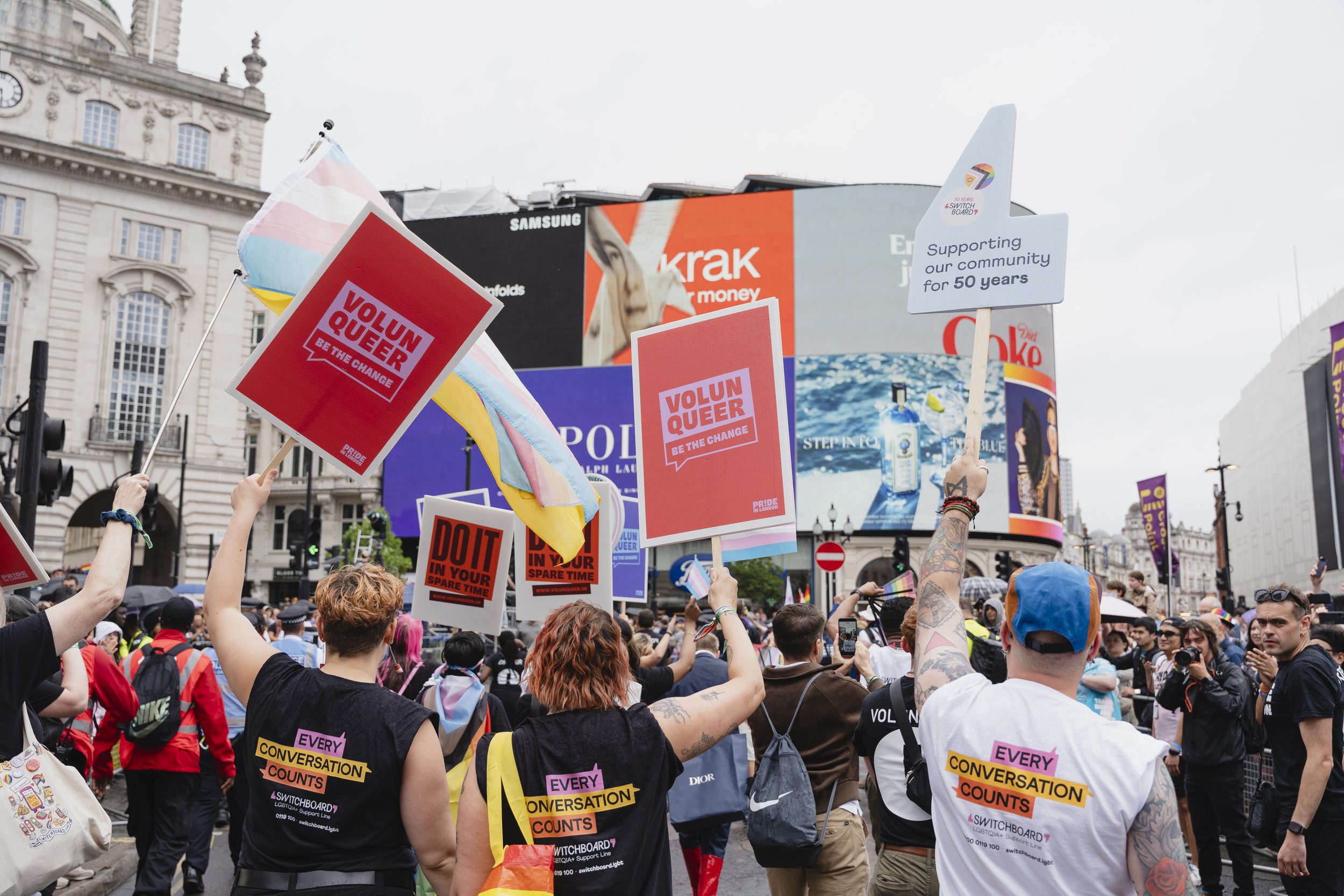 A view from behind a group of marchers in Piccadilly Circus during the Pride in London parade. Several people hold up red signs that read "VOLUN-QUEER: BE THE CHANGE,"