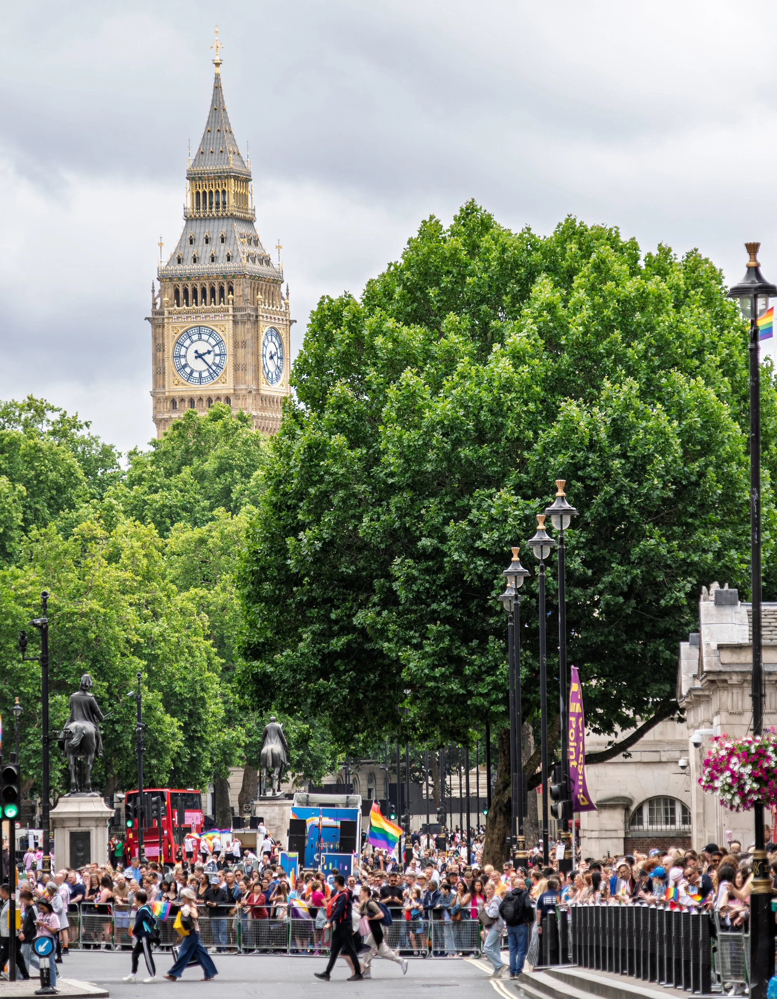 A wide vertical shot of the Pride in London parade route with the Elizabeth Tower (Big Ben) rising above a canopy of lush green trees in the background. In the foreground, a dense crowd of spectators lines the street behind metal barriers