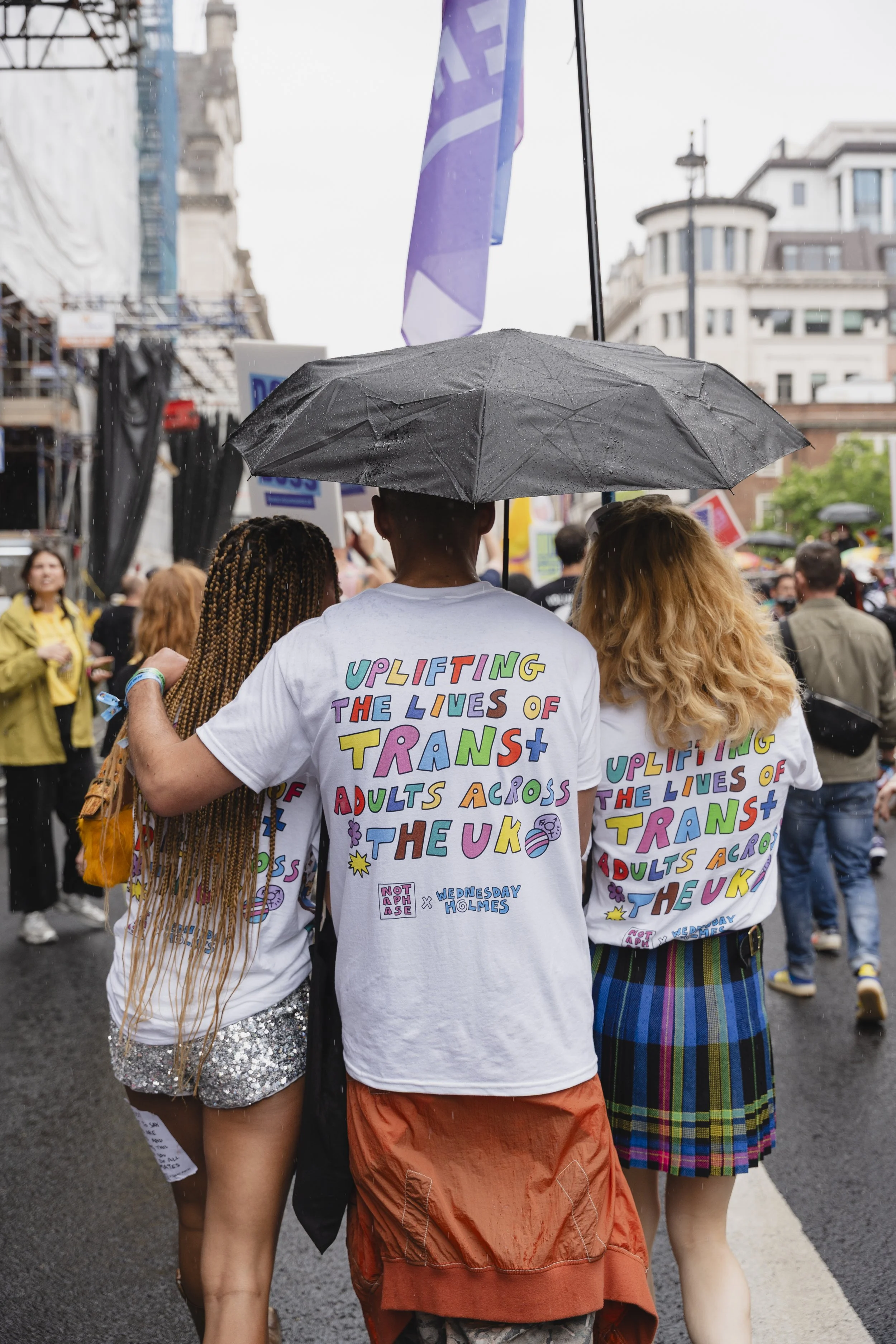A view from behind of three individuals walking together in the Pride in London parade under a black umbrella during a light rain. They are wearing matching white t-shirts with colorful, hand-drawn style lettering