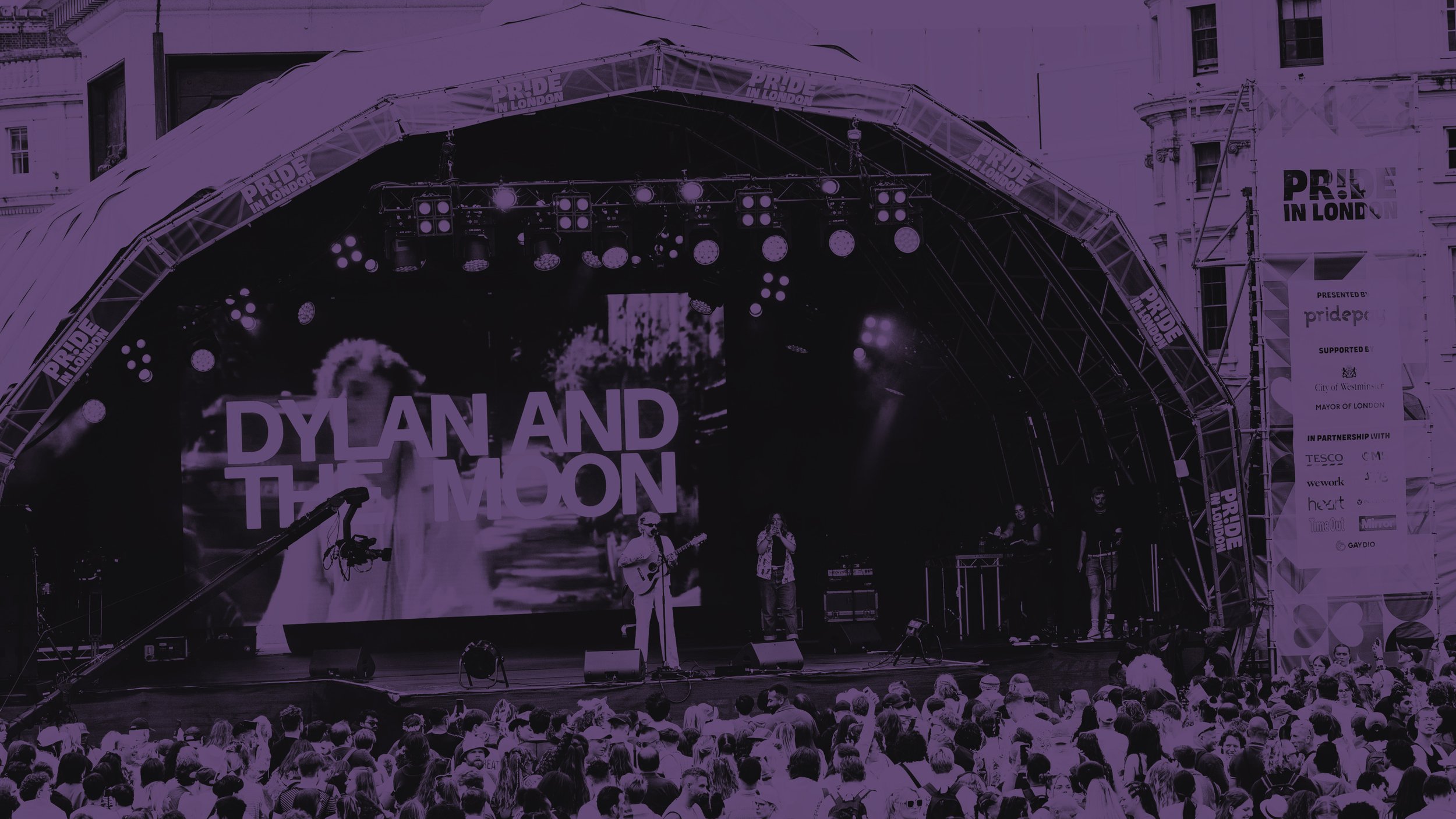 A purple-tinted wide shot of a Pride in London stage. Musicians perform for a large crowd; a screen displays "DYLAN AND THE MOON." Event banners and city buildings are visible in the background.