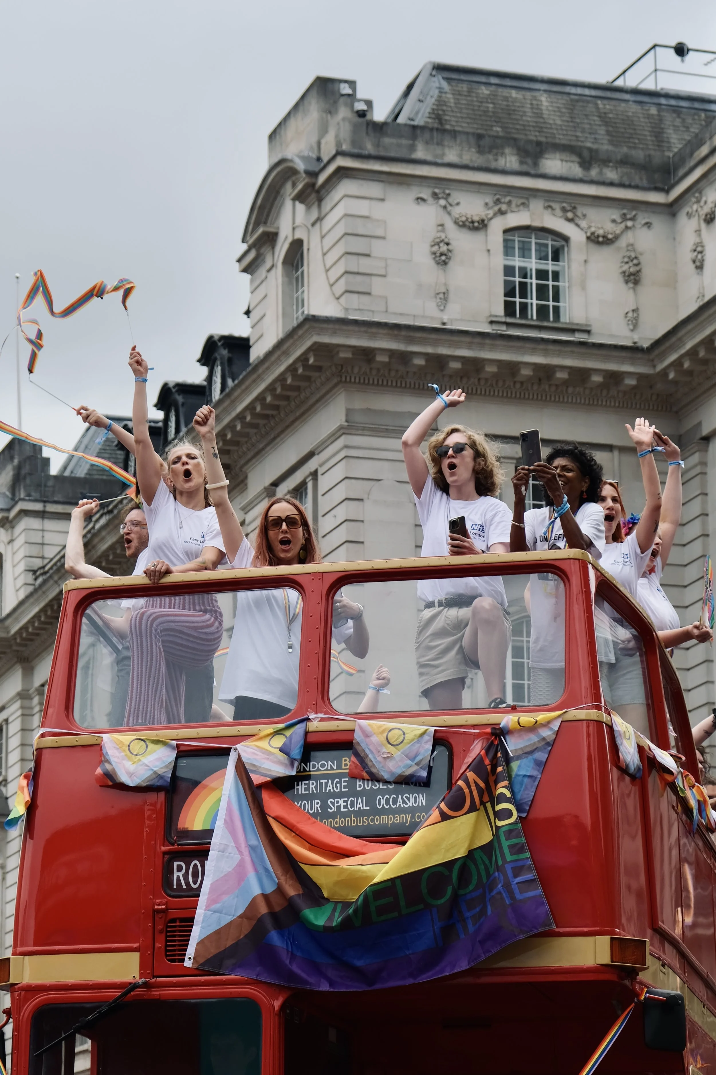 A group of people celebrate from the open-top deck of a vintage red double-decker bus during the Pride in London parade. The participants, many wearing white t-shirts, are cheering, waving rainbow ribbons, and taking photos