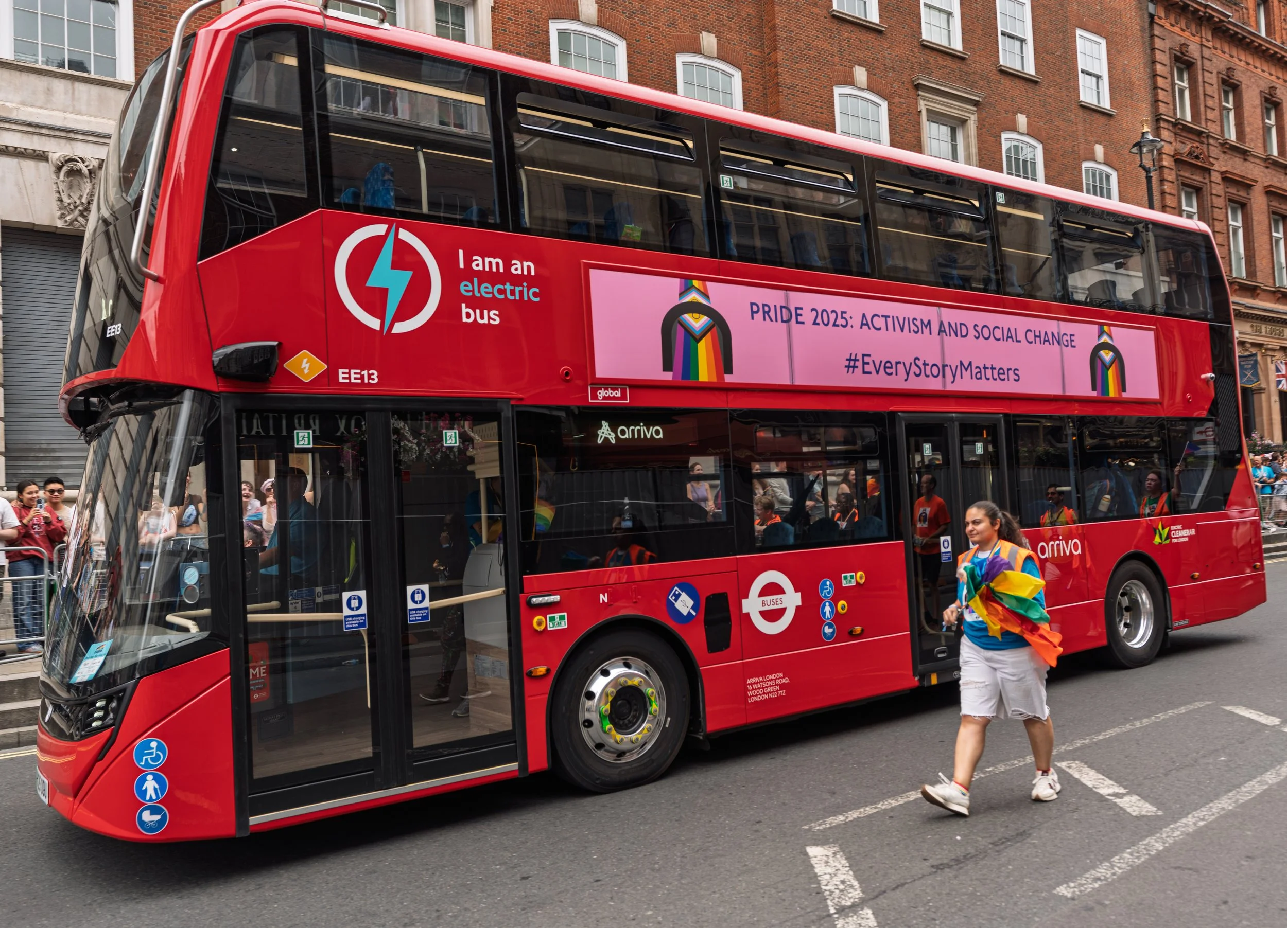 A bright red electric double-decker bus drives through a London street as part of the Pride parade. The side of the bus features a large pink banner that reads "PRIDE 2025: ACTIVISM AND SOCIAL CHANGE #EveryStoryMatters."