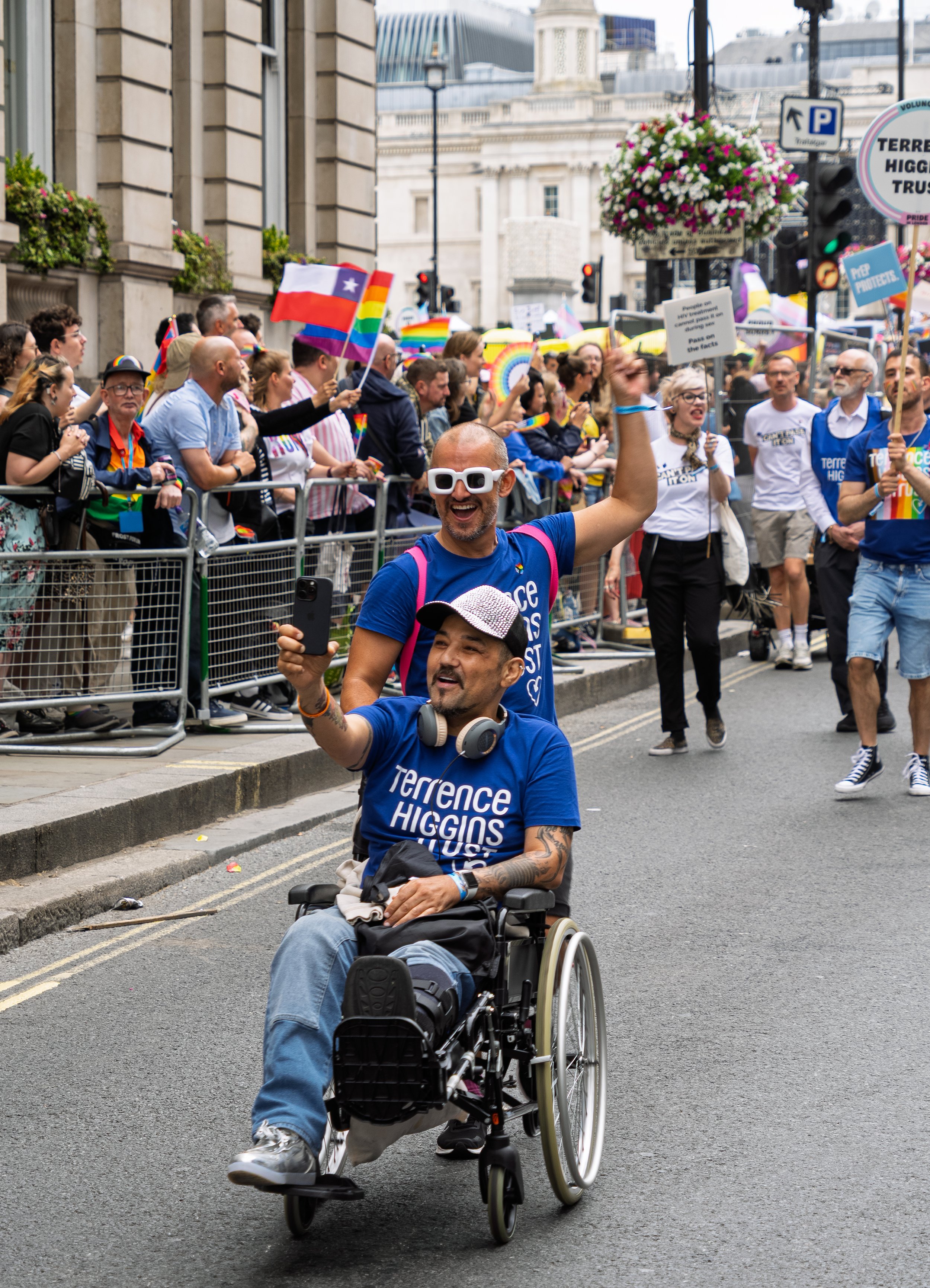 A smiling man in a wheelchair takes a selfie with a cheering friend at Pride in London. Both wear blue Terrence Higgins Trust shirts while marching past crowds lining a historic city street.