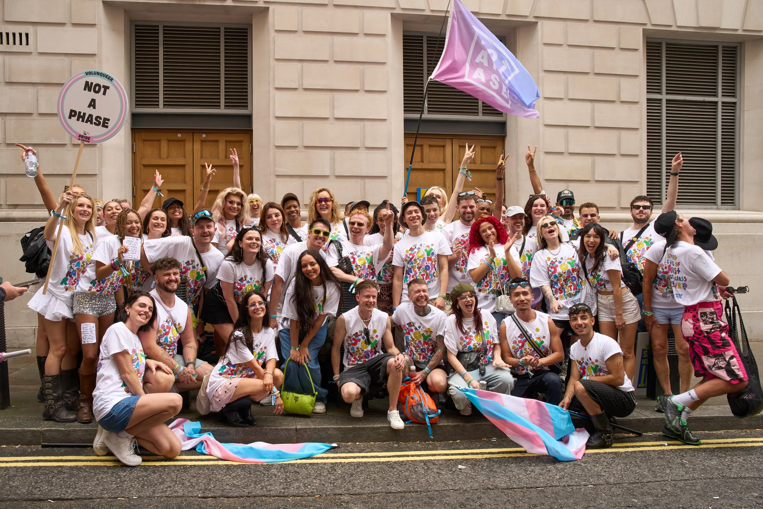 Group photo of Pride volunteers in matching white T shirts, cheering outside a building.