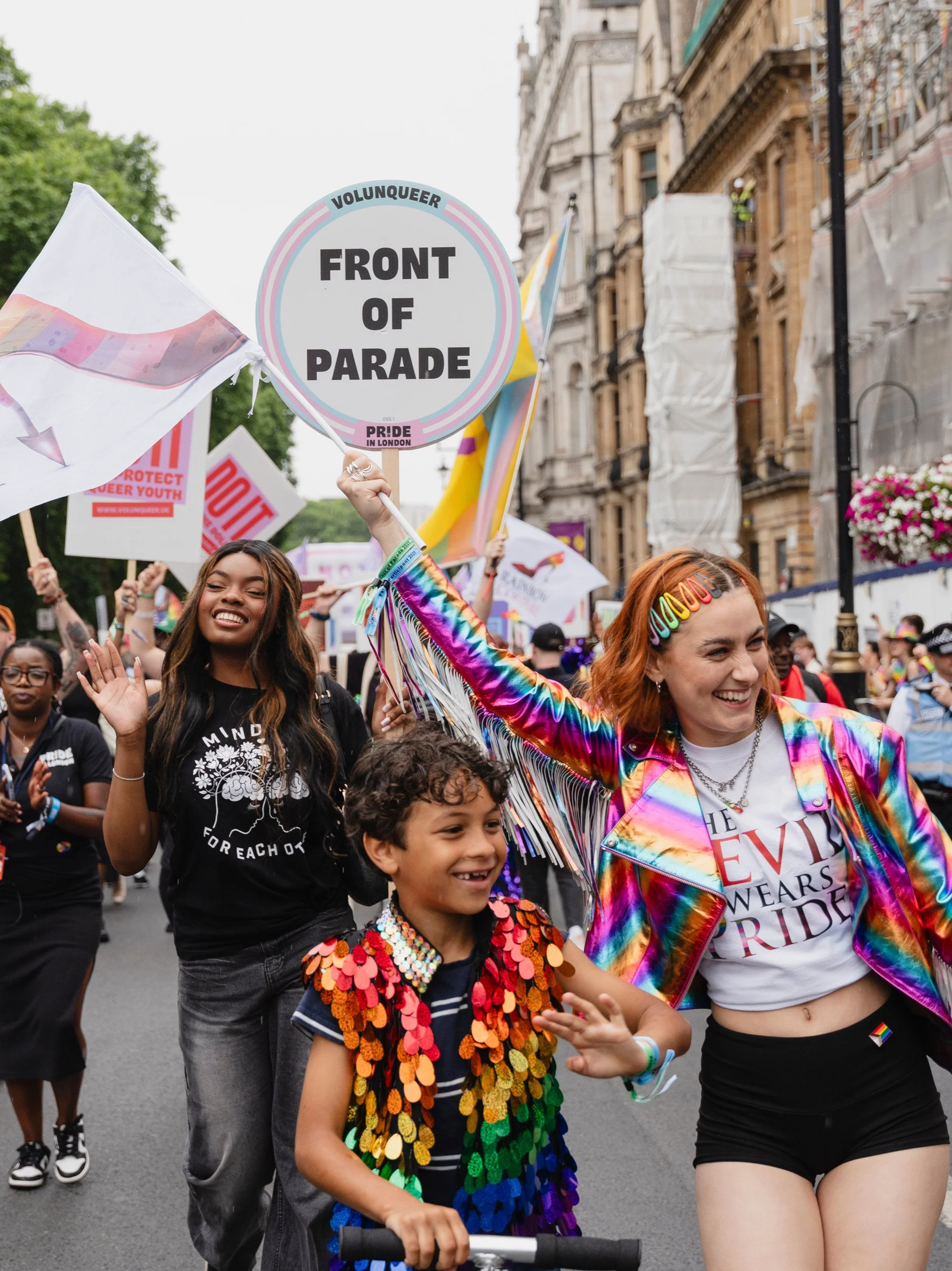 A joyful group of participants marches at the front of the Pride in London parade. In the center, a child wearing a colorful sequined vest waves while riding a scooter.