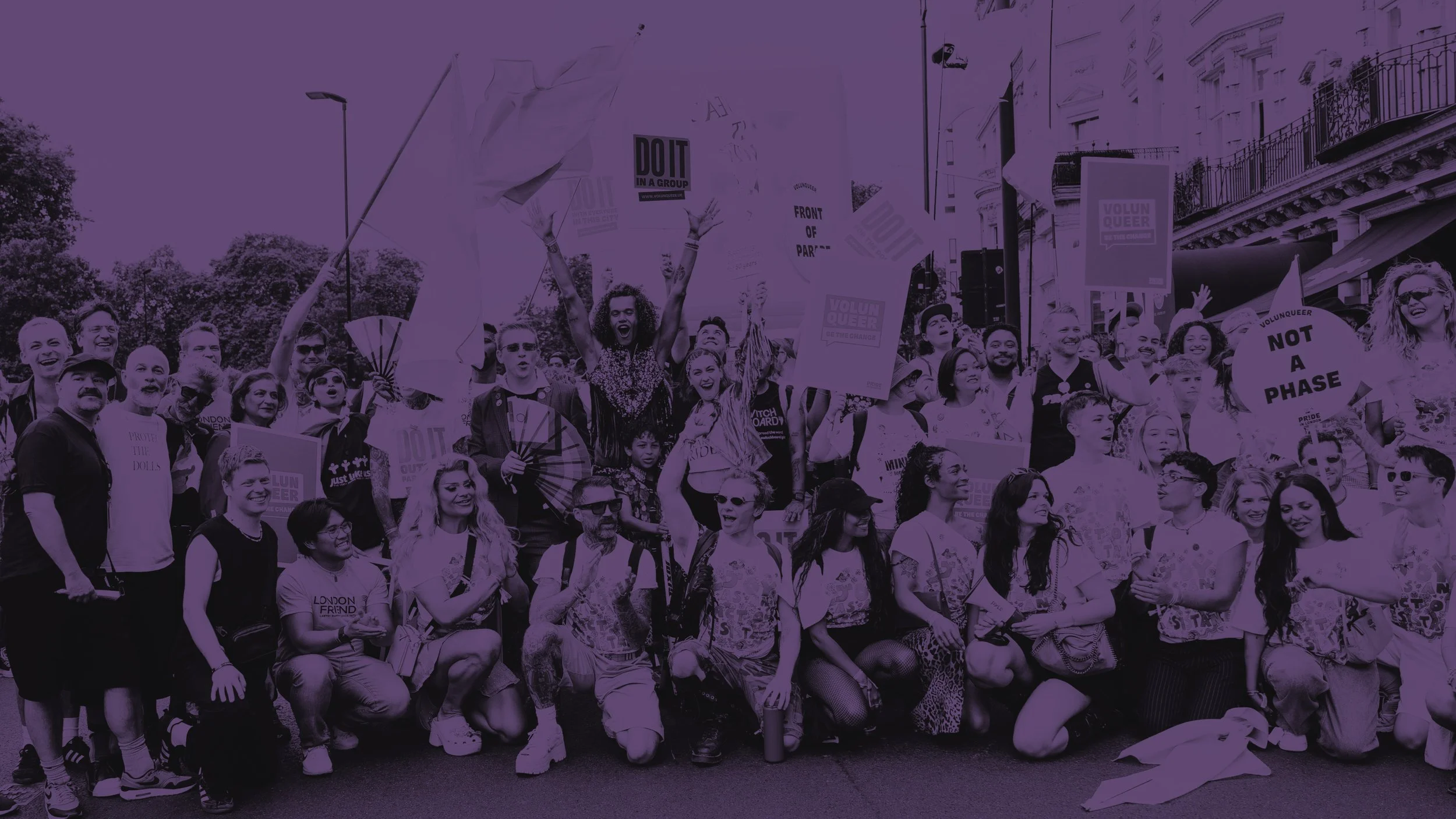 A large group celebrating at a Pride event, shown in a purple tint. People are smiling and holding signs like "NOT A PHASE" and "VOLUNQUEER" on a city street. The atmosphere is joyful and communal.