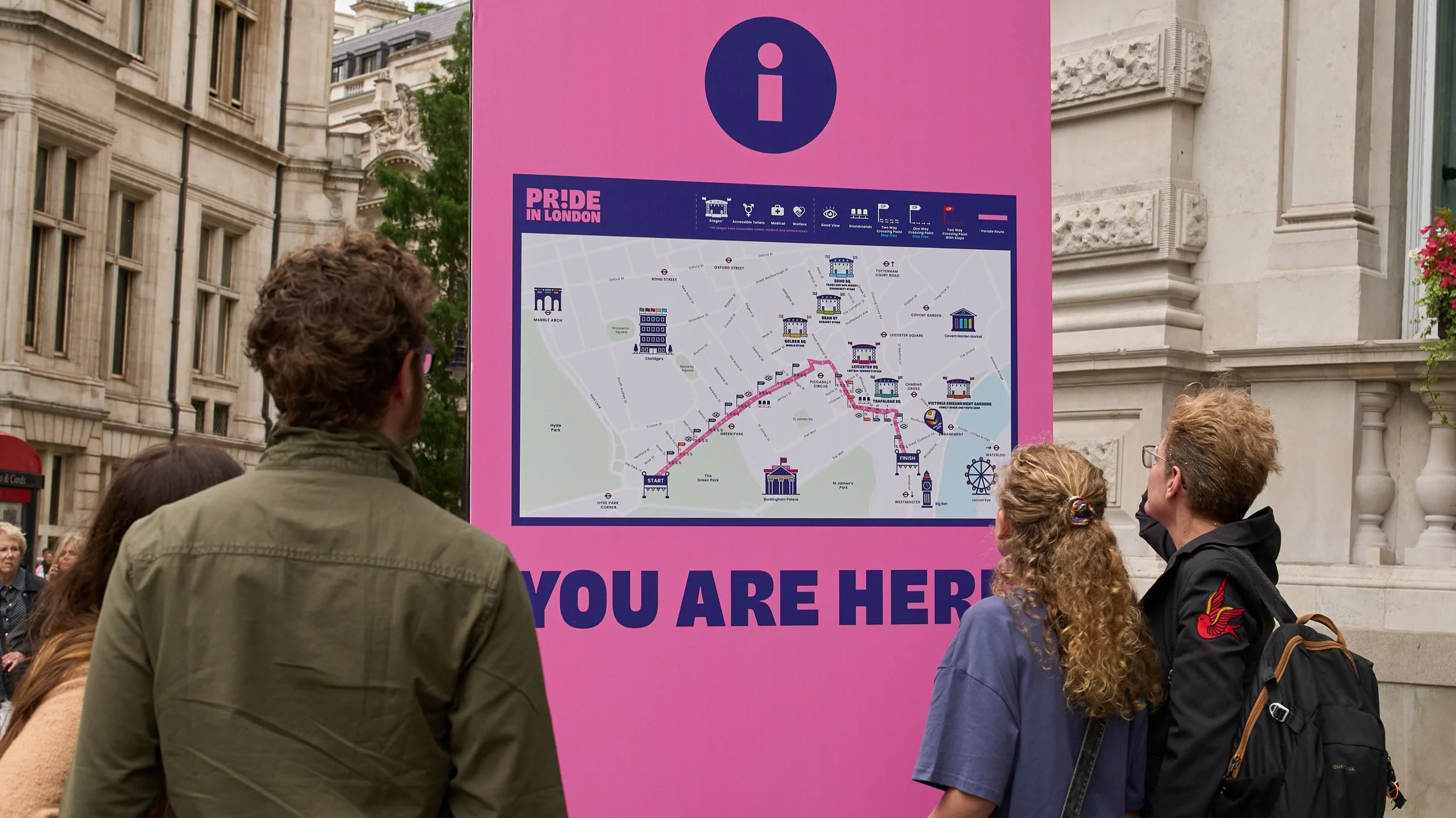 A group of attendees consults a large, vibrant pink information pillar in Central London. The pillar features a detailed map of the Pride in London parade route, highlighting various stage locations and facility icons.