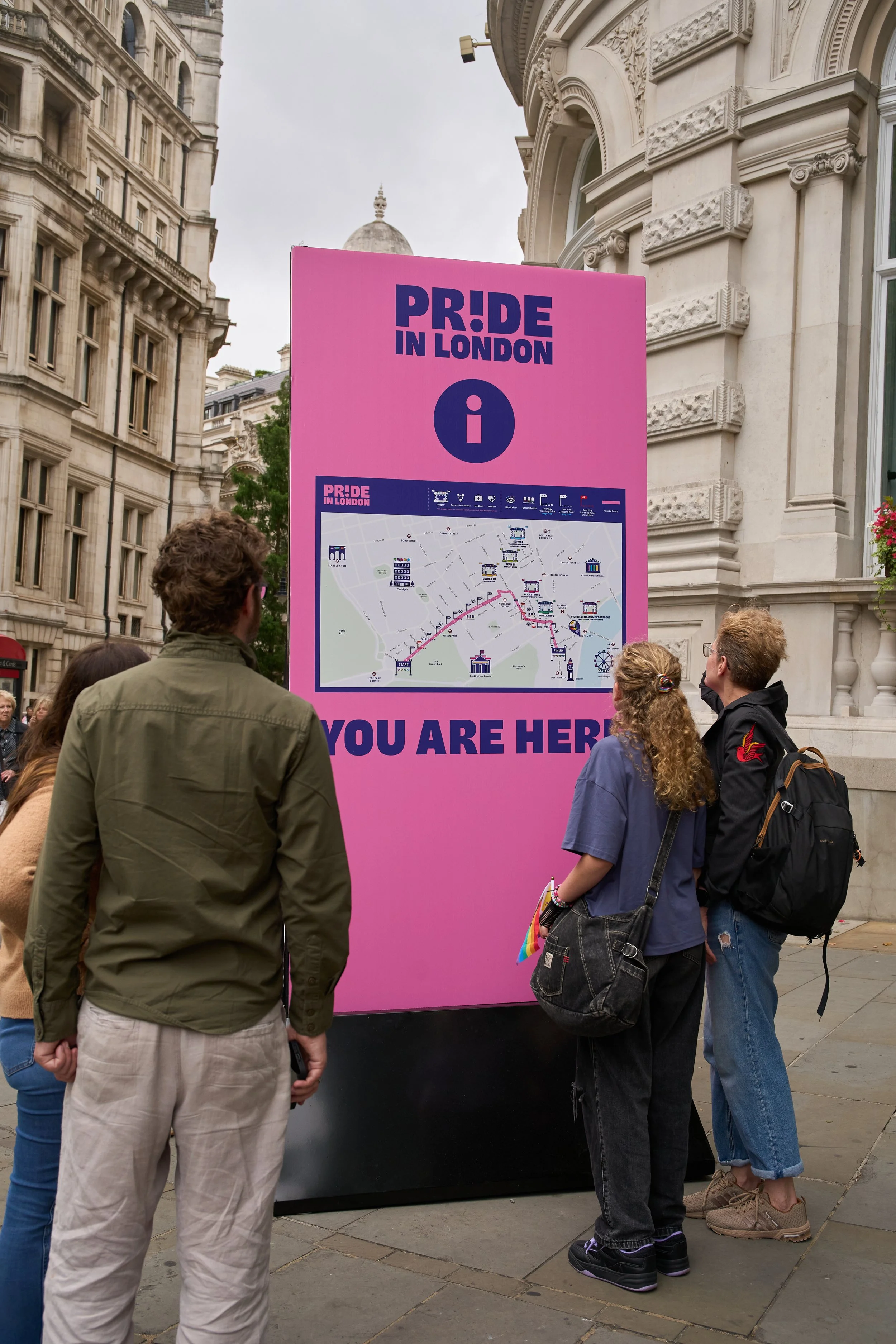 A tall pink Pride in London wayfinding board featuring a detailed event map and "YOU ARE HERE" text, with attendees using it for navigation.