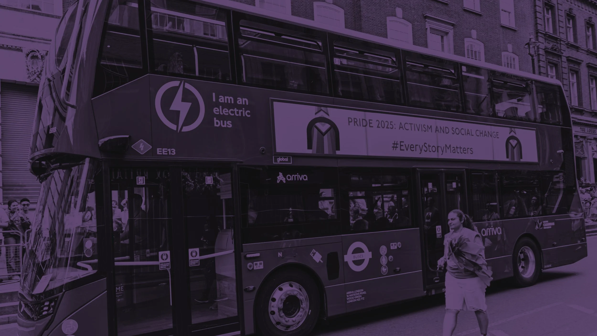 A purple-tinted photo of a double-decker electric bus in London. A banner on the side reads "PRIDE 2025: ACTIVISM AND SOCIAL CHANGE #EveryStoryMatters." A person walks past the bus in the foreground.