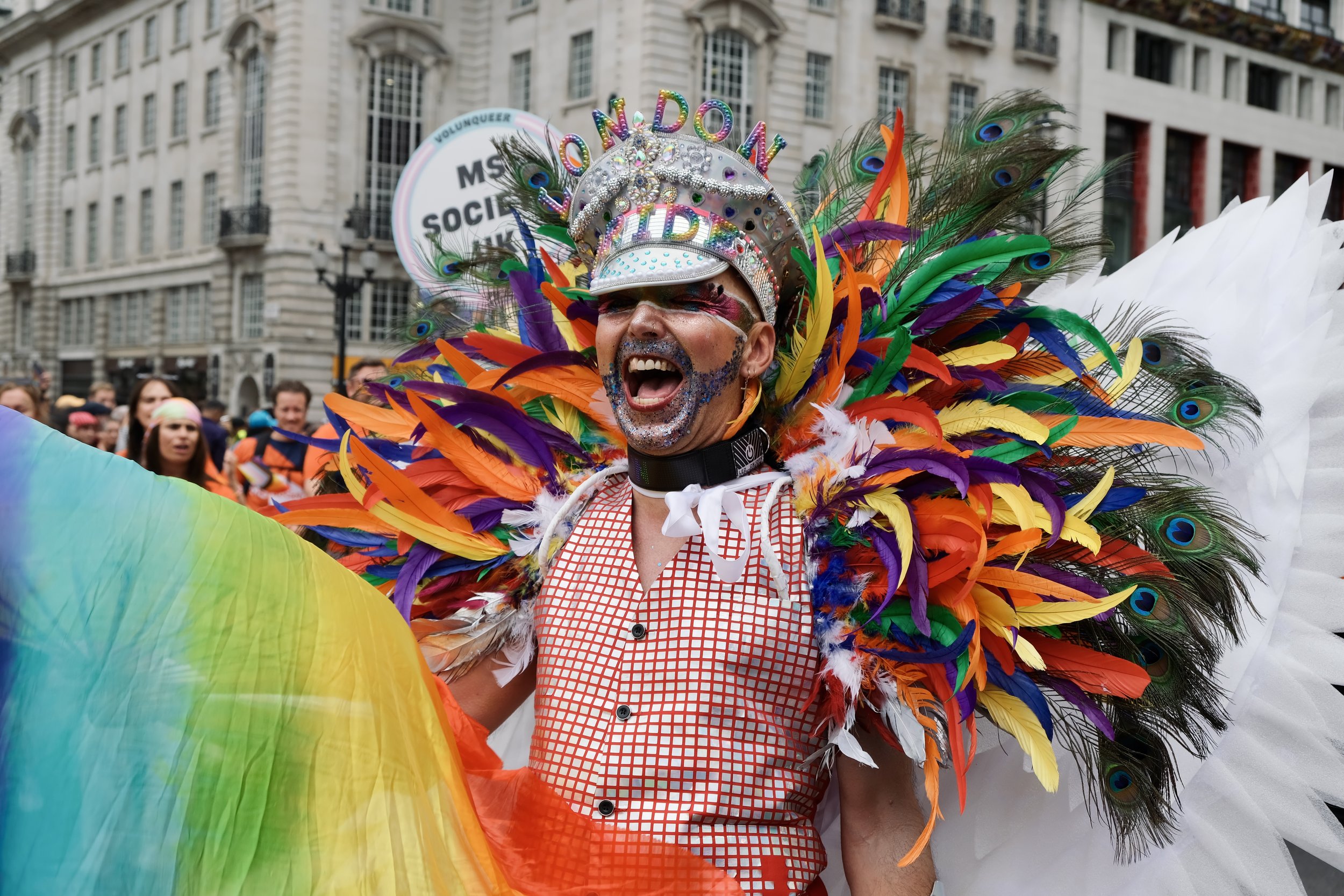 A person celebrates at the Pride in London parade wearing an elaborate, colorful costume featuring a massive collar of rainbow and peacock feathers. They wear a silver-jeweled captain's hat that reads "LONDON PRIDE"