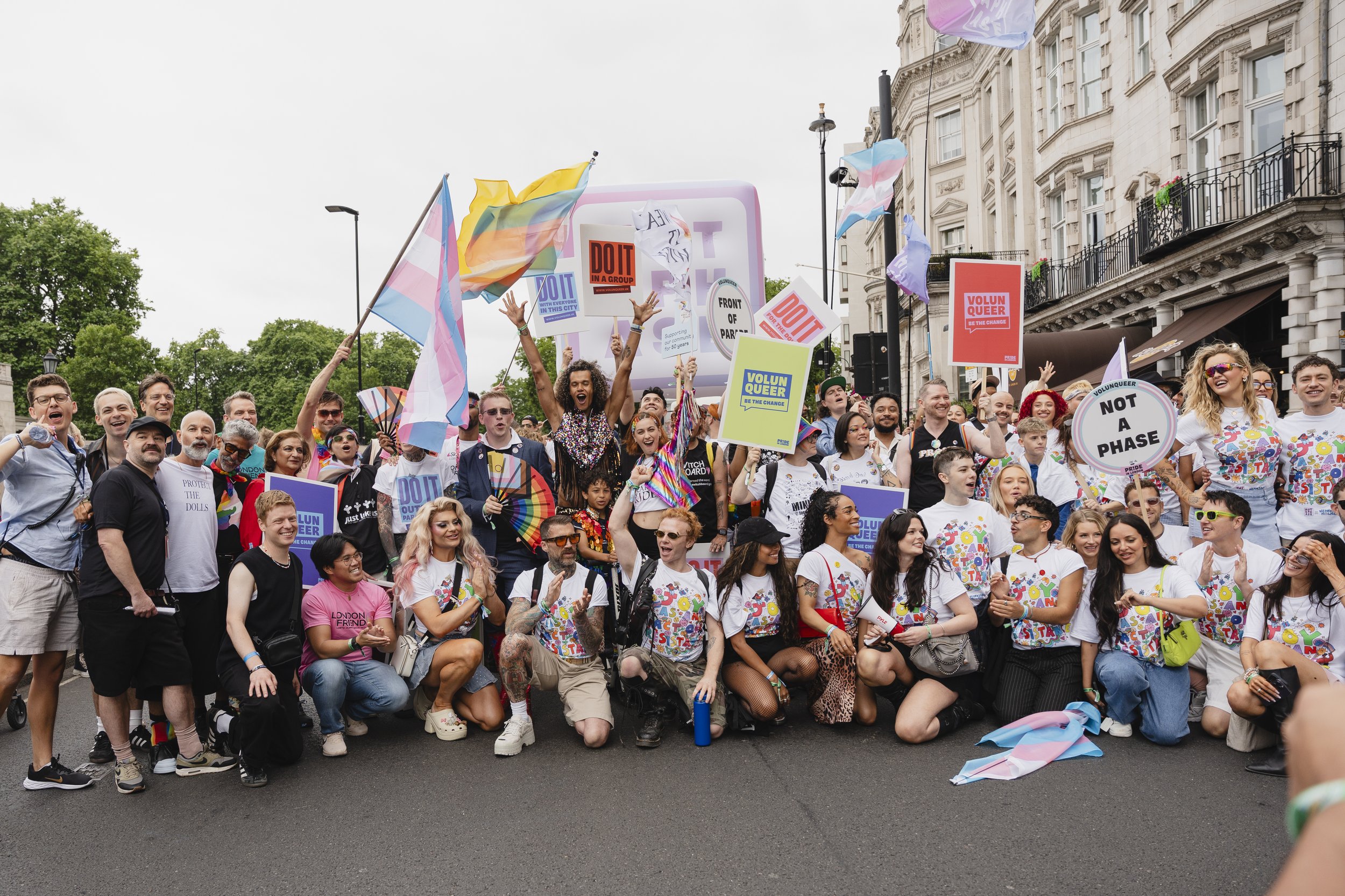 A large, joyful group of Pride in London participants poses together on a city street. Many wear "Joy is Resistance" t-shirts and carry colorful signs with slogans like "Volun-queer," "Not a Phase," and "Front of Parade."