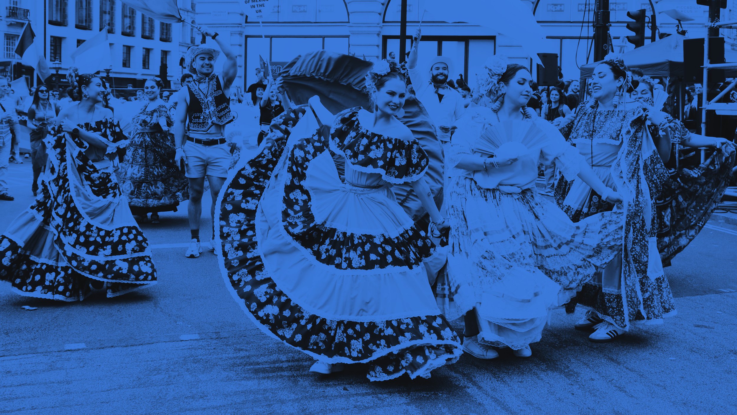 Dancers in vibrant, flowing traditional dresses perform at Pride in London. Behind them, people wave the Mexican and Transgender Pride flags against a backdrop of historic city buildings.