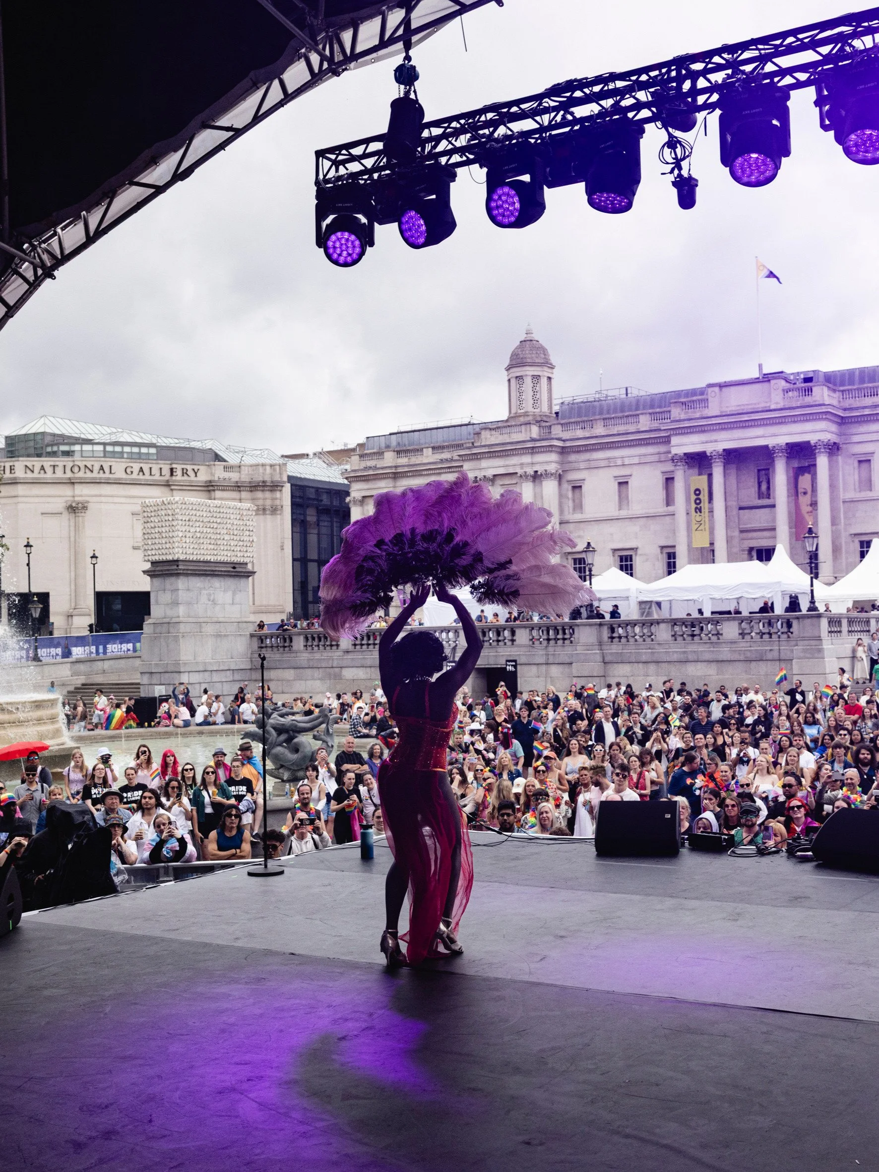 A wide shot from the back of a stage in Trafalgar Square, looking out over a massive crowd during Pride in London. A performer in a red costume and sheer skirt stands center stage with their back to the camera,