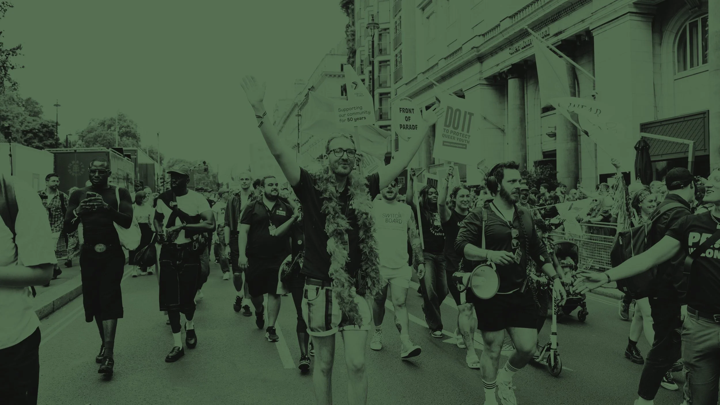 A green-tinted photo of a Pride parade. A person in a feather boa cheers with arms raised among a crowd holding "FRONT OF PARADE" and "DO IT" signs. The mood is celebratory and high-energy.