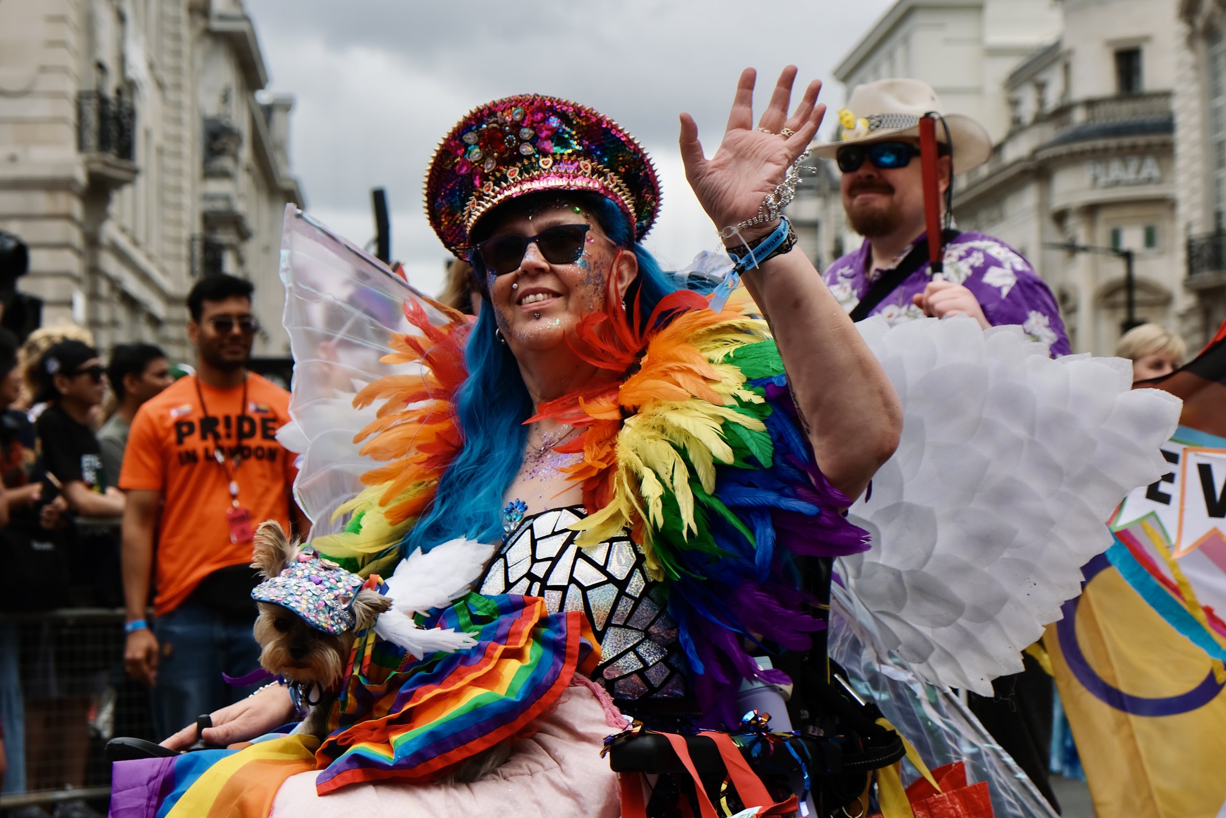 A smiling person in a wheelchair participates in the Pride in London parade, waving to the crowd. They are wearing a vibrant rainbow feather collar, a sequined hat, and white wings, with a small dog in a matching rainbow outfit on their lap.