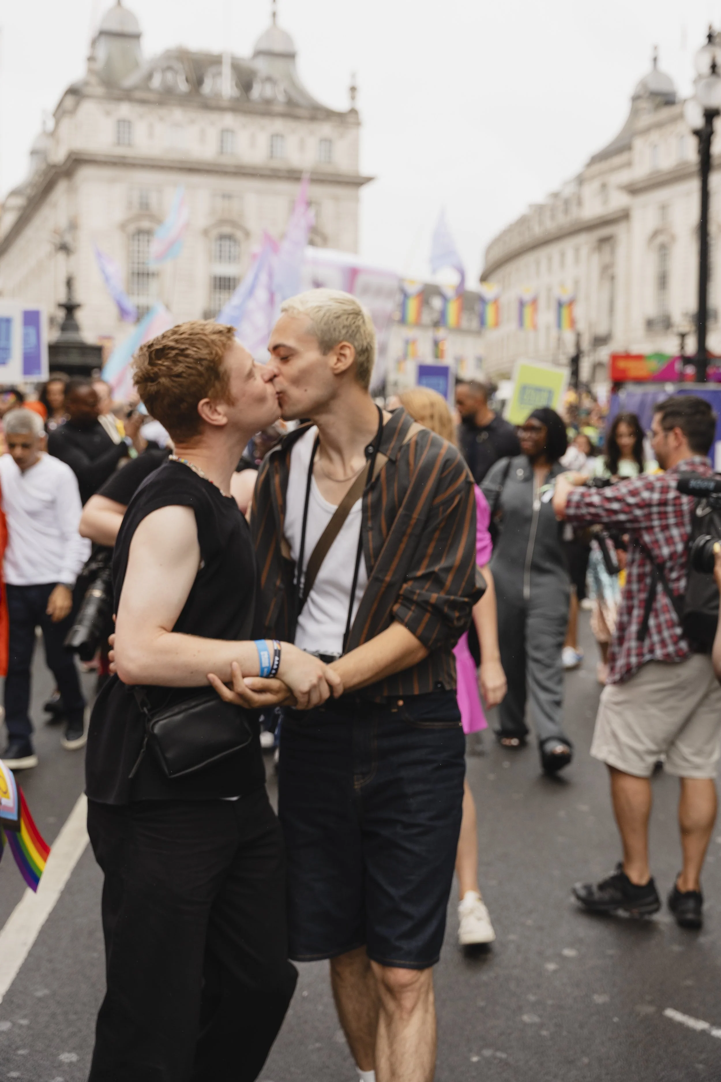 Two people share a romantic kiss in the middle of a busy street during the Pride in London parade. One person wears a black sleeveless top and cross-body bag, while the other wears a striped shirt over a white tank top and denim shorts.