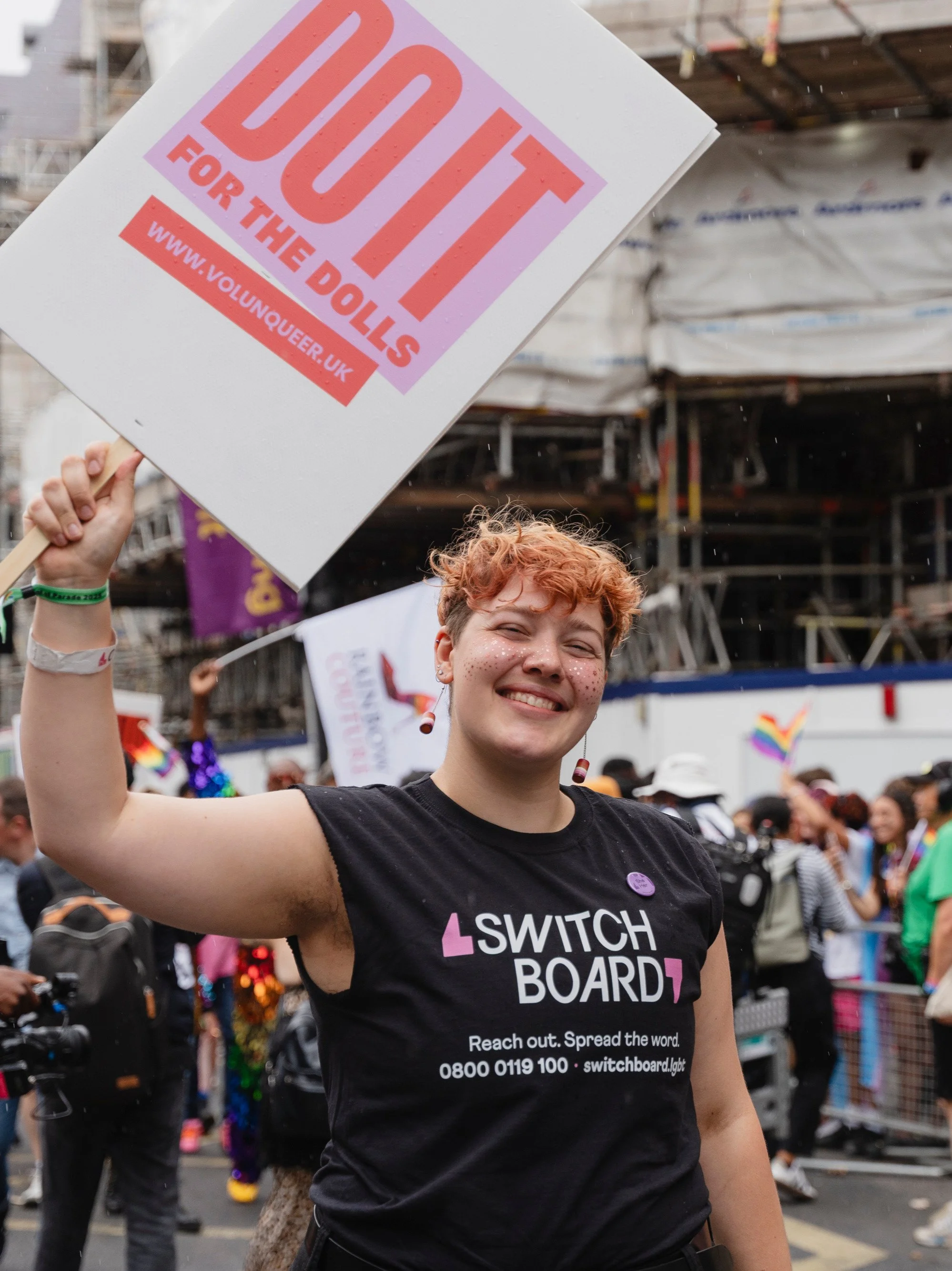 A smiling participant with short orange hair and glitter on their face holds up a large placard that says "DO IT FOR THE DOLLS" in bold pink letters.