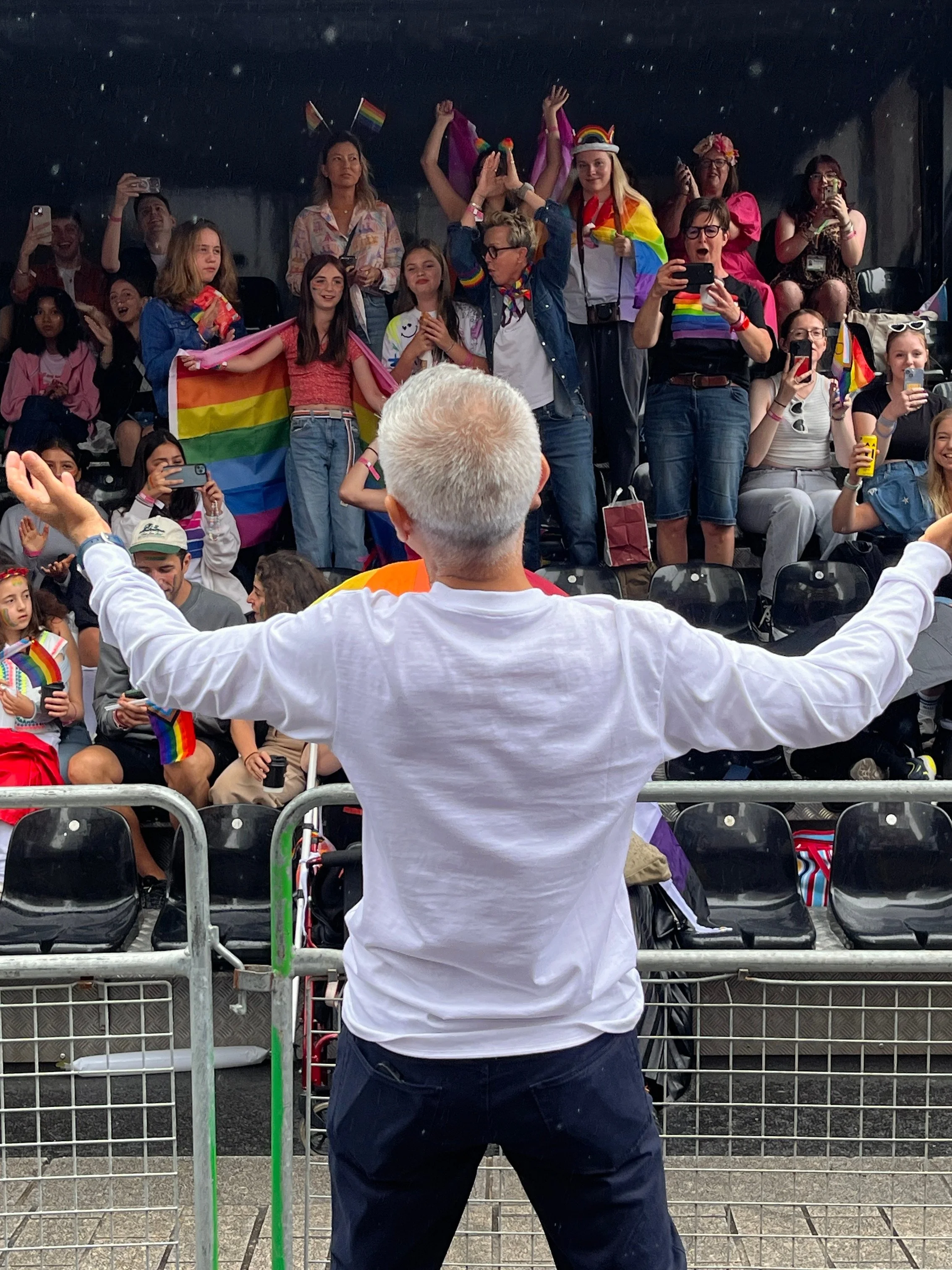 A view from behind a person in a white long-sleeved shirt with their arms outstretched toward a tiered grandstand. The grandstand is filled with a diverse crowd of people cheering, taking photos, and waving rainbow flags.