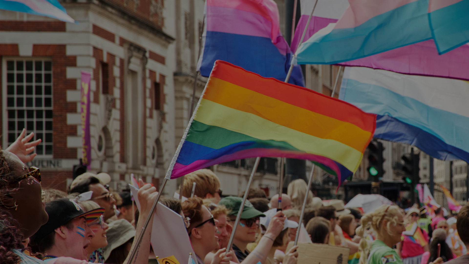 A vibrant Pride parade crowd waving colorful LGBTQIA+ pride, bisexual, and trans flags along a city street lined with historic brick buildings.