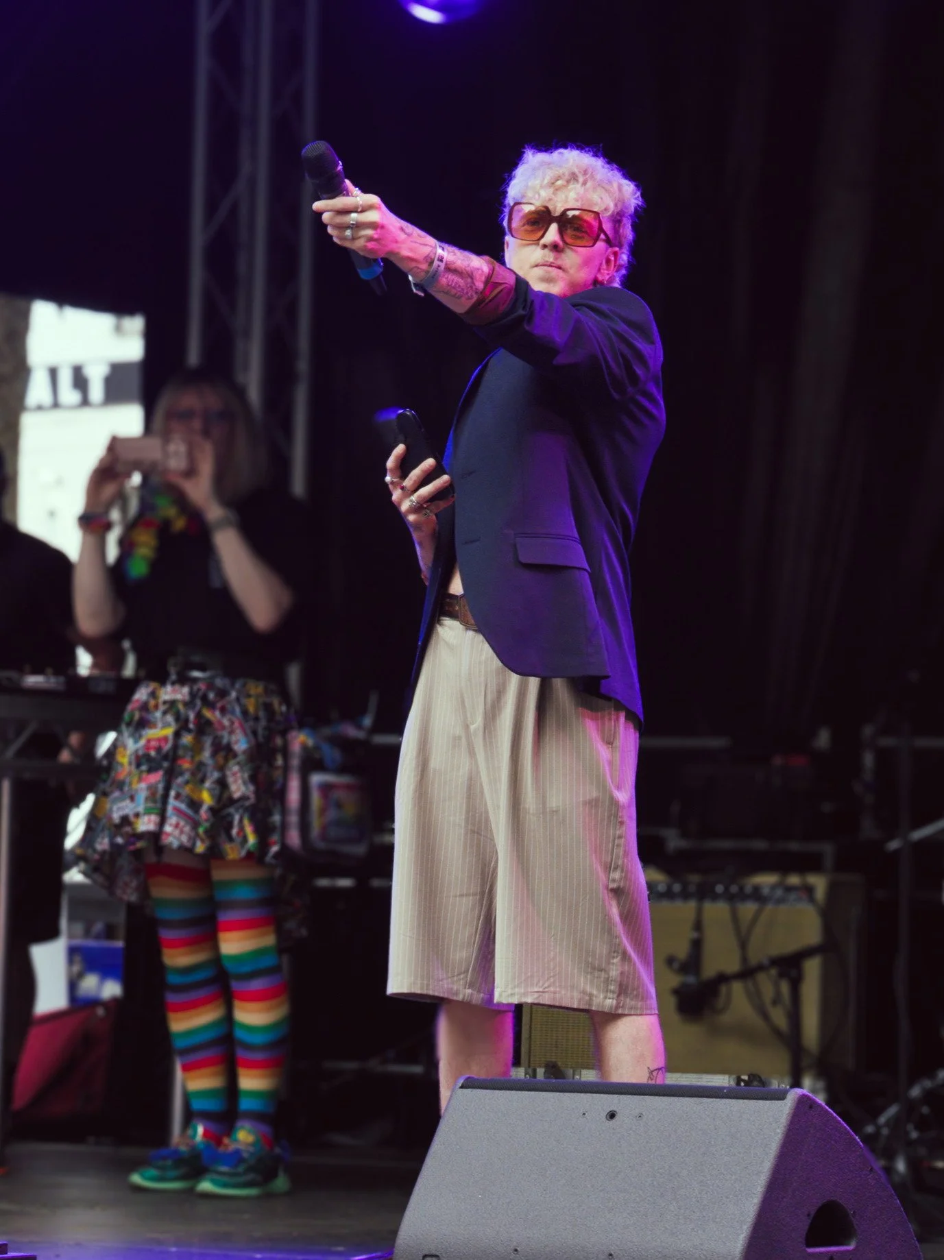 A performer with blonde curly hair and oversized orange sunglasses stands on a stage during Pride in London. They are wearing a dark navy blazer over pinstriped tan shorts, holding a microphone out toward the audience in a commanding pose.