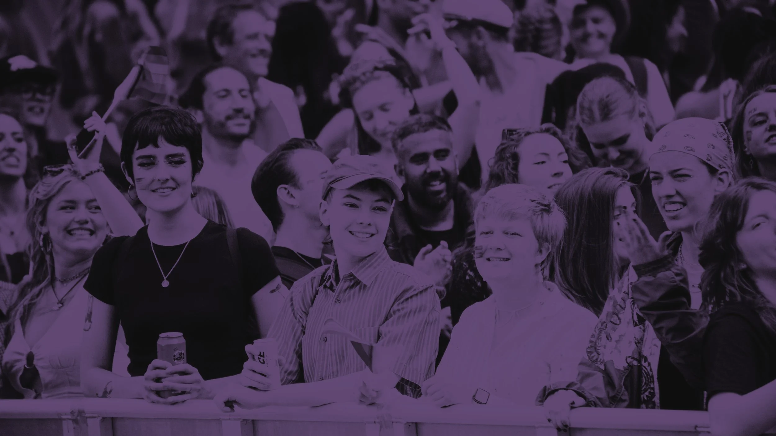 A purple-tinted close-up of a smiling crowd behind a security barrier at a Pride event. People are cheering and holding drinks, capturing a joyful, communal atmosphere at an outdoor festival.
