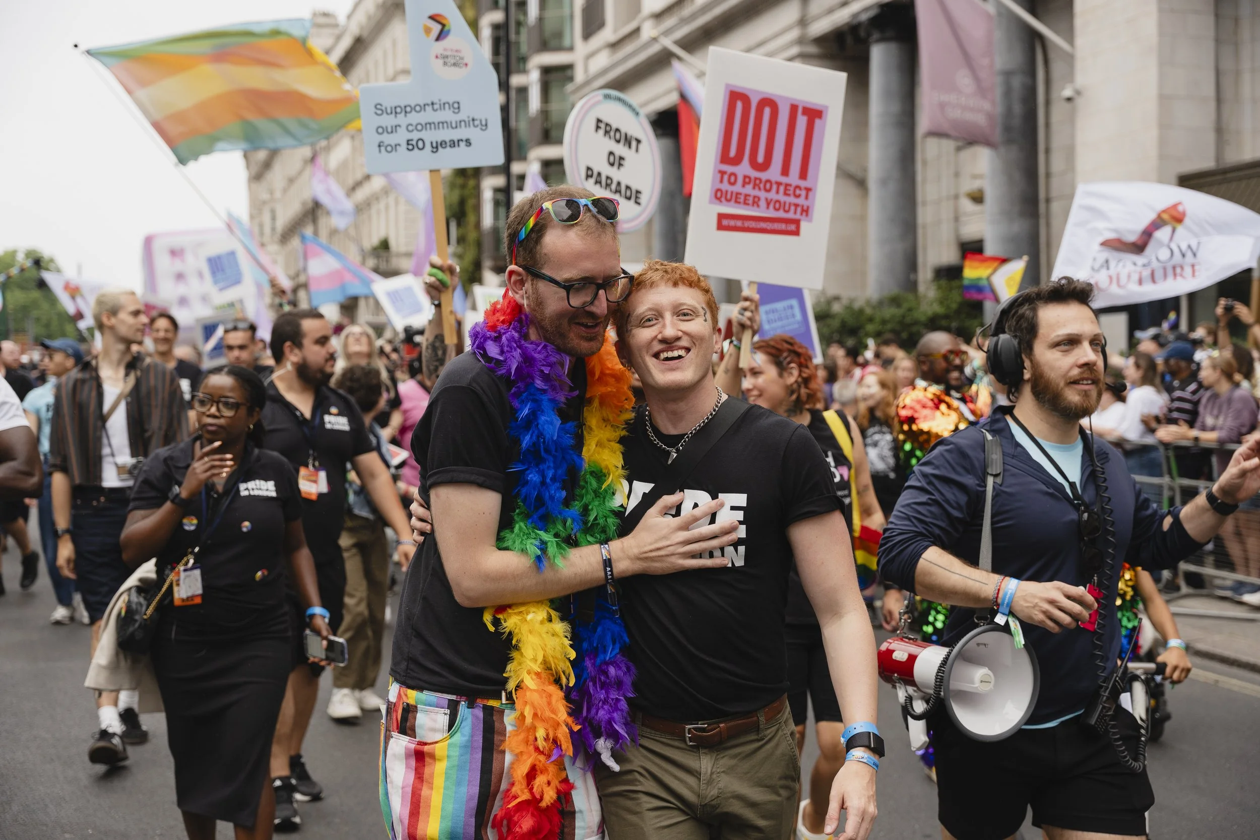 Two men embrace and smile warmly during the Pride in London parade. One wears a rainbow feather boa and rainbow-striped trousers, the other wears a black "Pride in London" t-shirt. They are surrounded by a diverse crowd under a city skyline.