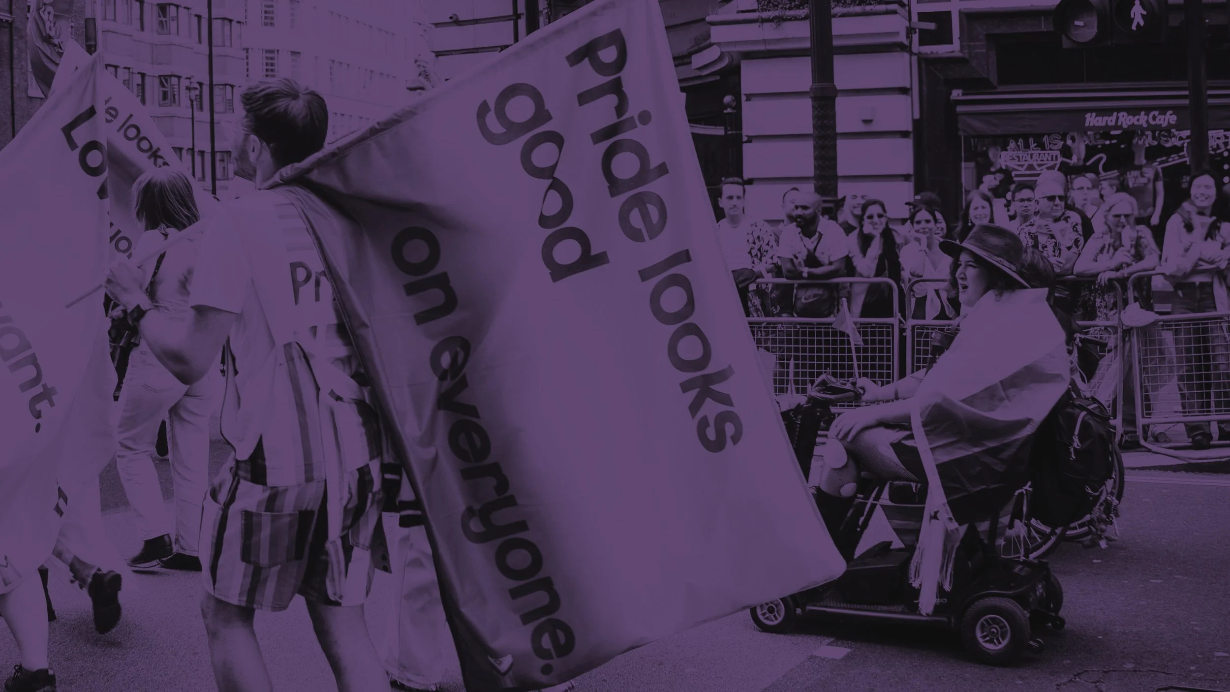 A purple-tinted photo from a Pride parade. A person marches with a large flag reading "Pride looks good on everyone." In the background, a person in a mobility scooter joins the joyful procession.