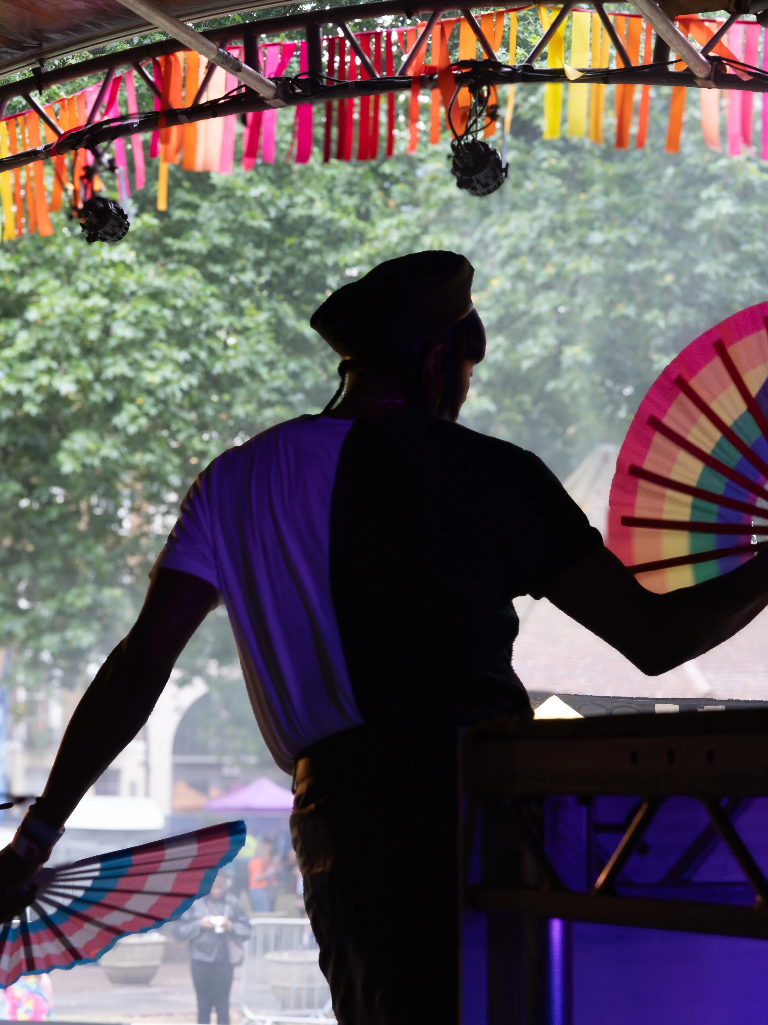 A silhouette of a performer on a stage at Pride in London, seen from behind. They are wearing a cap and holding two large folding fans—one with rainbow stripes and the other with transgender flag colors.