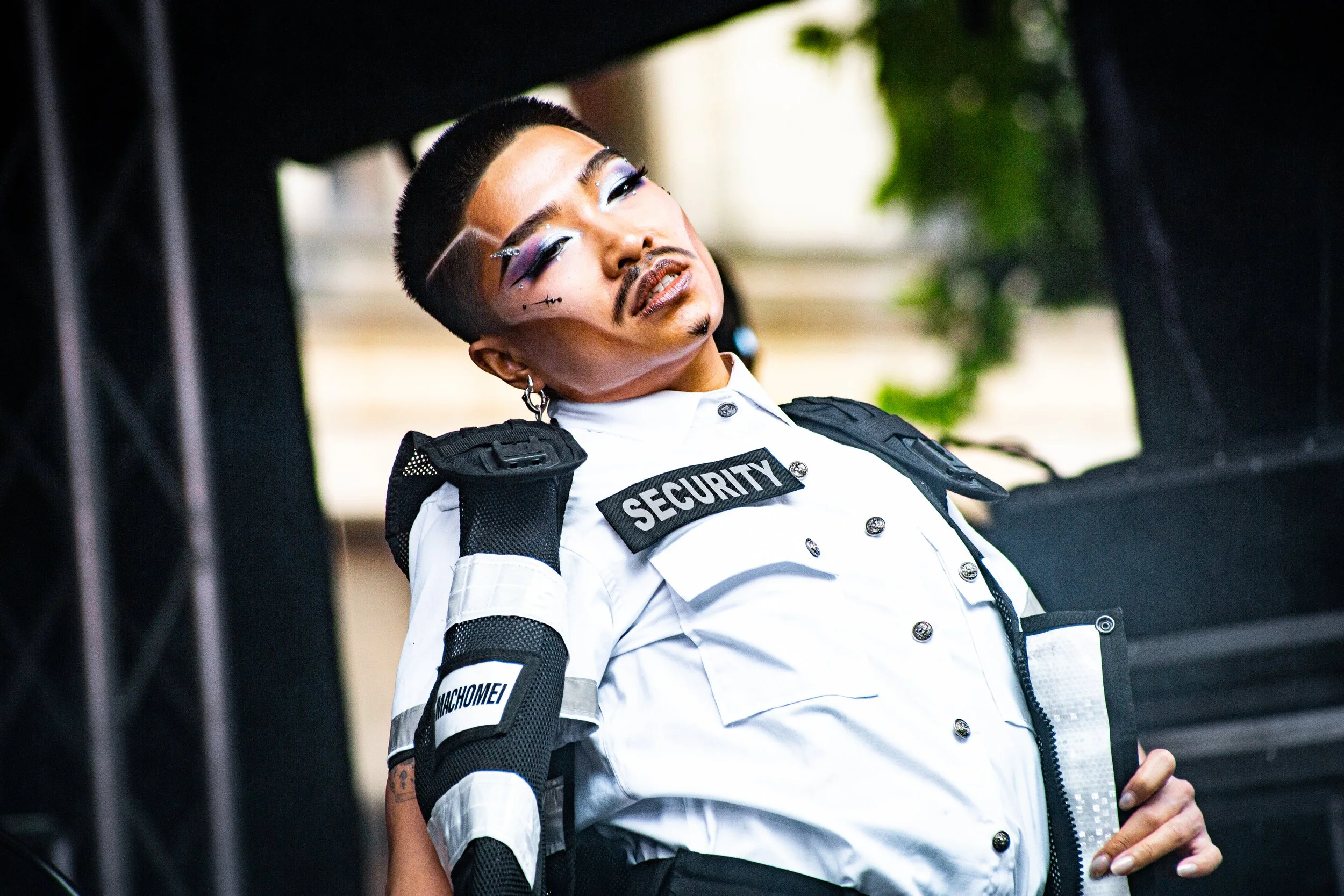 A performer in striking, glittery eye makeup and a stylized mustache poses on stage wearing a white uniform shirt with a "SECURITY" patch, body armor pads, and a short buzzcut with shaved lines.