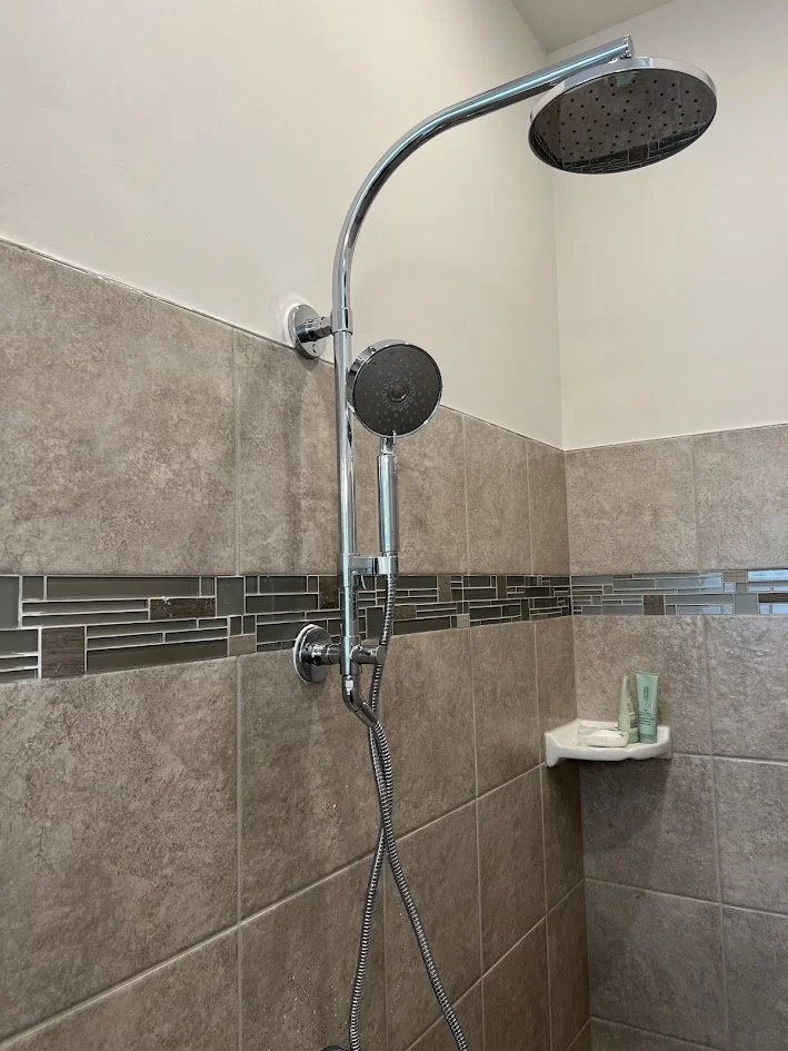 Shower with a chrome showerhead, handheld shower, beige and gray tile walls, and a small corner shelf with toiletries.