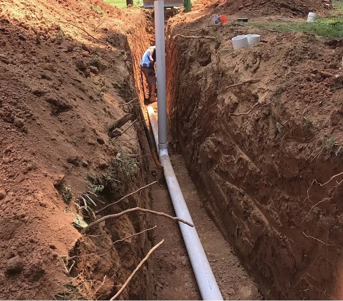 A person working in a deep trench installing or repairing a large gray pipe, with dirt walls on both sides and construction tools and materials nearby.