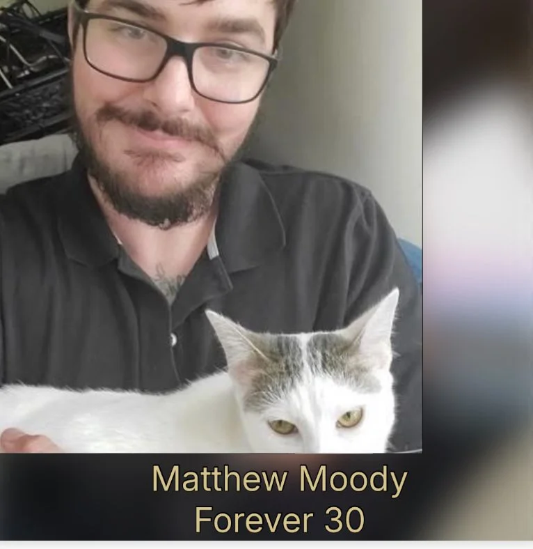 Photo of a man with glasses and a beard taking a selfie with a white and gray cat. There is text at the bottom reading, "Matthew Moody Forever 30."