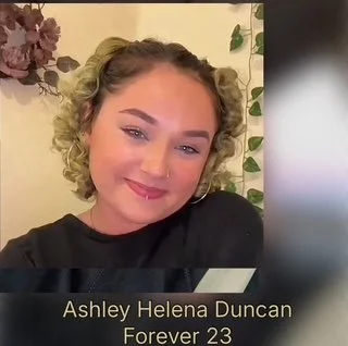 A young woman with blonde, curly hair taking a selfie. She is smiling softly and wearing a black top. The background includes plants and floral decorations. Text overlay reads: 'Ashley Helena Duncan Forever 23'.