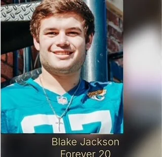 Young man smiling, wearing a blue sports jersey and a necklace, outdoors with a playground structure in the background, a sign reading 'Blake Jackson Forever 20' at the bottom.