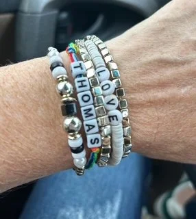 A person wearing multiple beaded bracelets on their wrist, including one with the words "I ❤️ LOVE THOMAS" and various colorful beads.