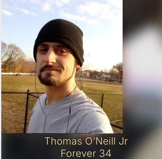 Portrait of a young man wearing a black beanie and a gray shirt, standing outdoors with a park and trees in the background. Text overlay reads: 'Thomas O'Neill Jr Forever 34'.