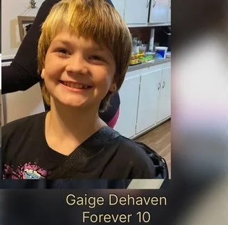 A smiling young boy with light brown hair in a black shirt, in a kitchen environment.