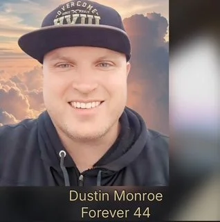 Young man smiling outdoors wearing a black baseball cap and hoodie, with a sunset sky in the background.