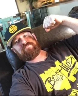 A man with a beard and a black baseball cap showing a yellow logo, sitting on a chair, flexing his right arm, wearing a black T-shirt with yellow text. He is inside a room with glass partitions.
