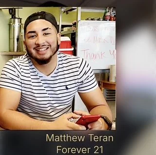 Young man smiling, wearing a striped shirt and black cap, sitting at a table with a red phone. Background shows shelves, a whiteboard with handwritten notes, and kitchen items.