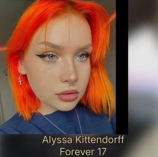 Close-up of a young woman with bright orange hair, piercing blue eyes, winged eyeliner, and multiple facial piercings, wearing a dark top and drop earrings.