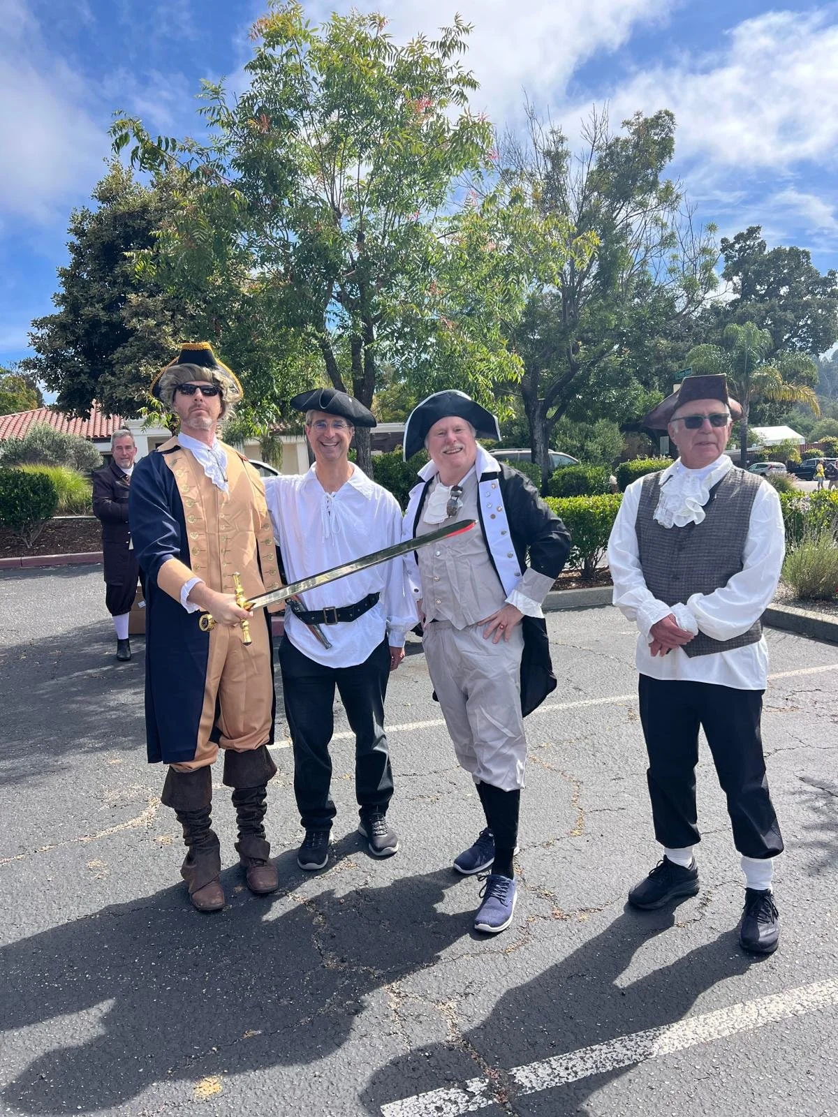 Four men dressed in 18th-century sailor and musketeer costumes standing in a parking lot under a tree, with a man in the background observing. The man on the left is holding a sword and wearing a tricorn hat, sunglasses, and period clothing. The othe